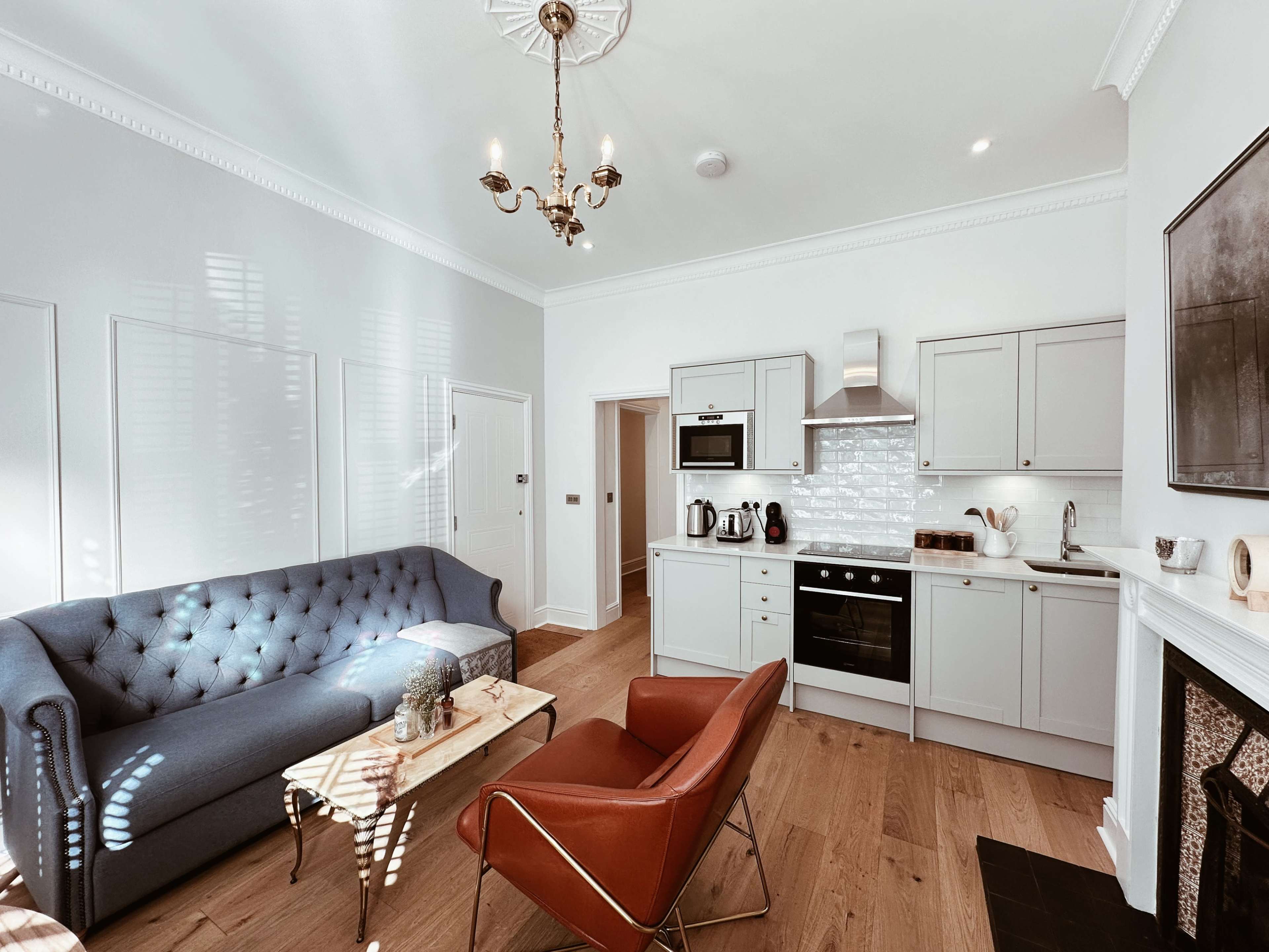 A modern living room with a blue tufted sofa, a red armchair, a wooden coffee table, and a kitchen area featuring light cabinetry and stainless steel appliances.