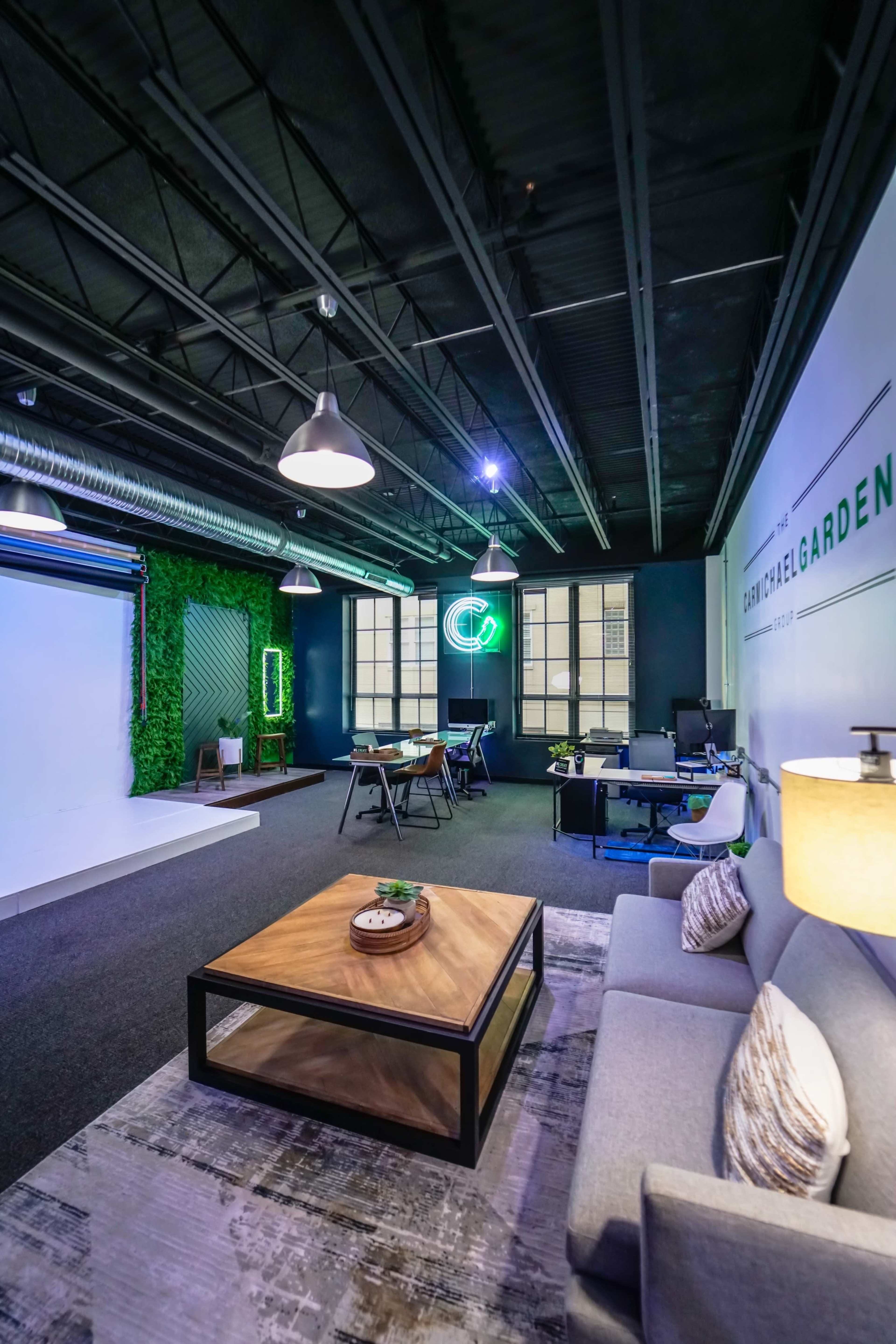A modern office space featuring a lounge area with a gray sofa and a wooden coffee table, alongside workstations and greenery, illuminated by overhead lighting.