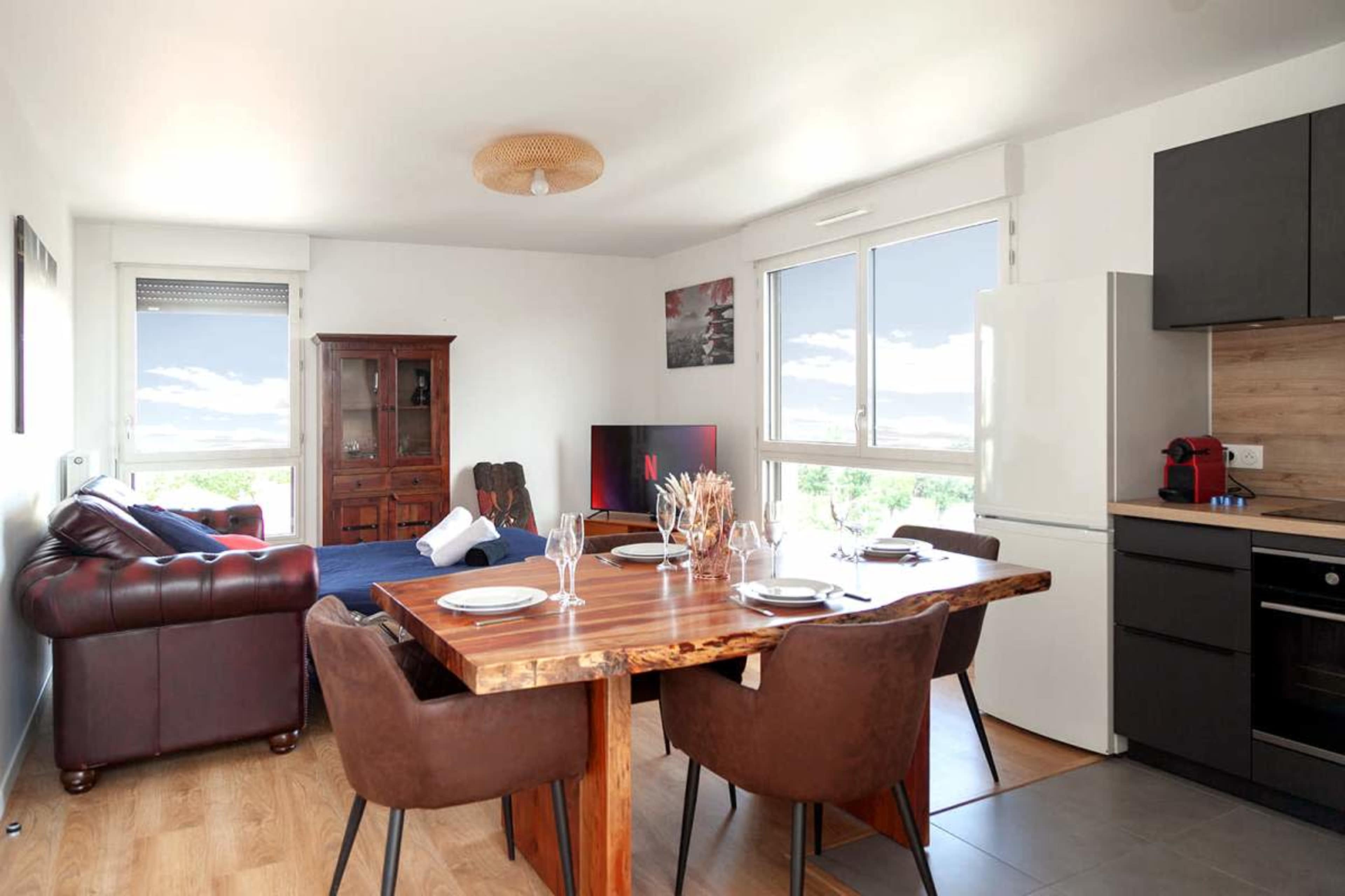 The image shows a modern open-concept living space featuring a dining table with chairs, a sofa, a television, and a kitchen area.