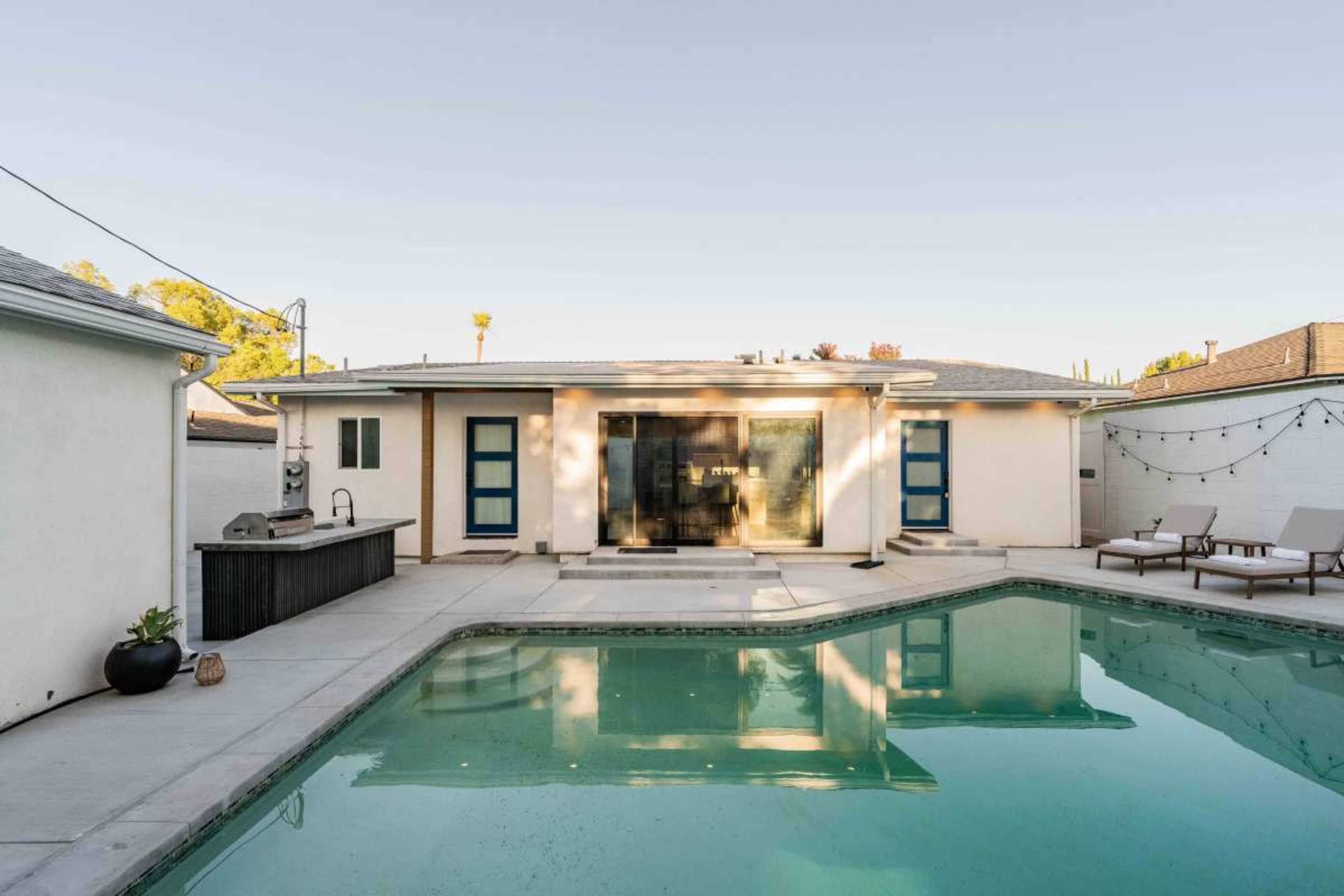 The image shows a modern backyard with a swimming pool, a patio area, and a house featuring large glass doors and blue accents.