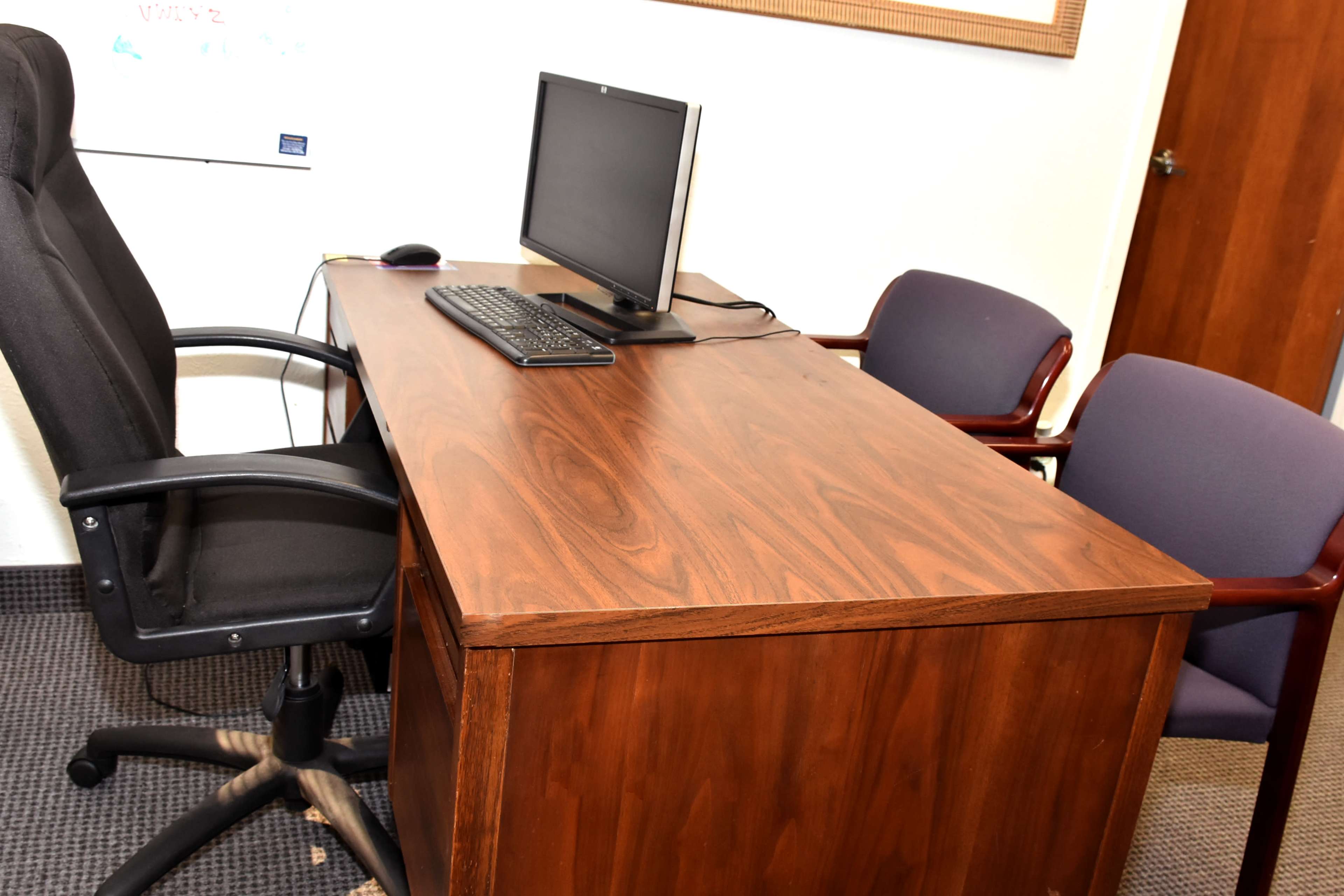 A wooden desk with a computer monitor, keyboard, and mouse is positioned in an office with two chairs nearby.