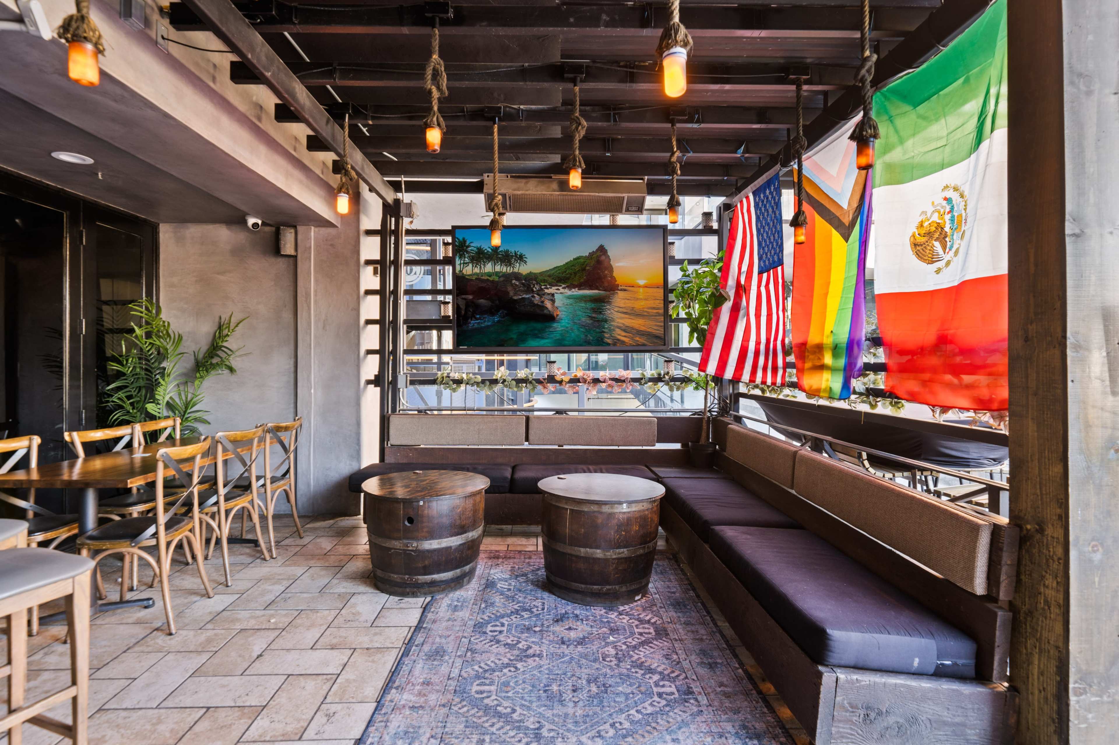 The image shows a semi-covered outdoor seating area with wooden barrels as tables, a patterned rug, and flags from various countries hanging on the walls.