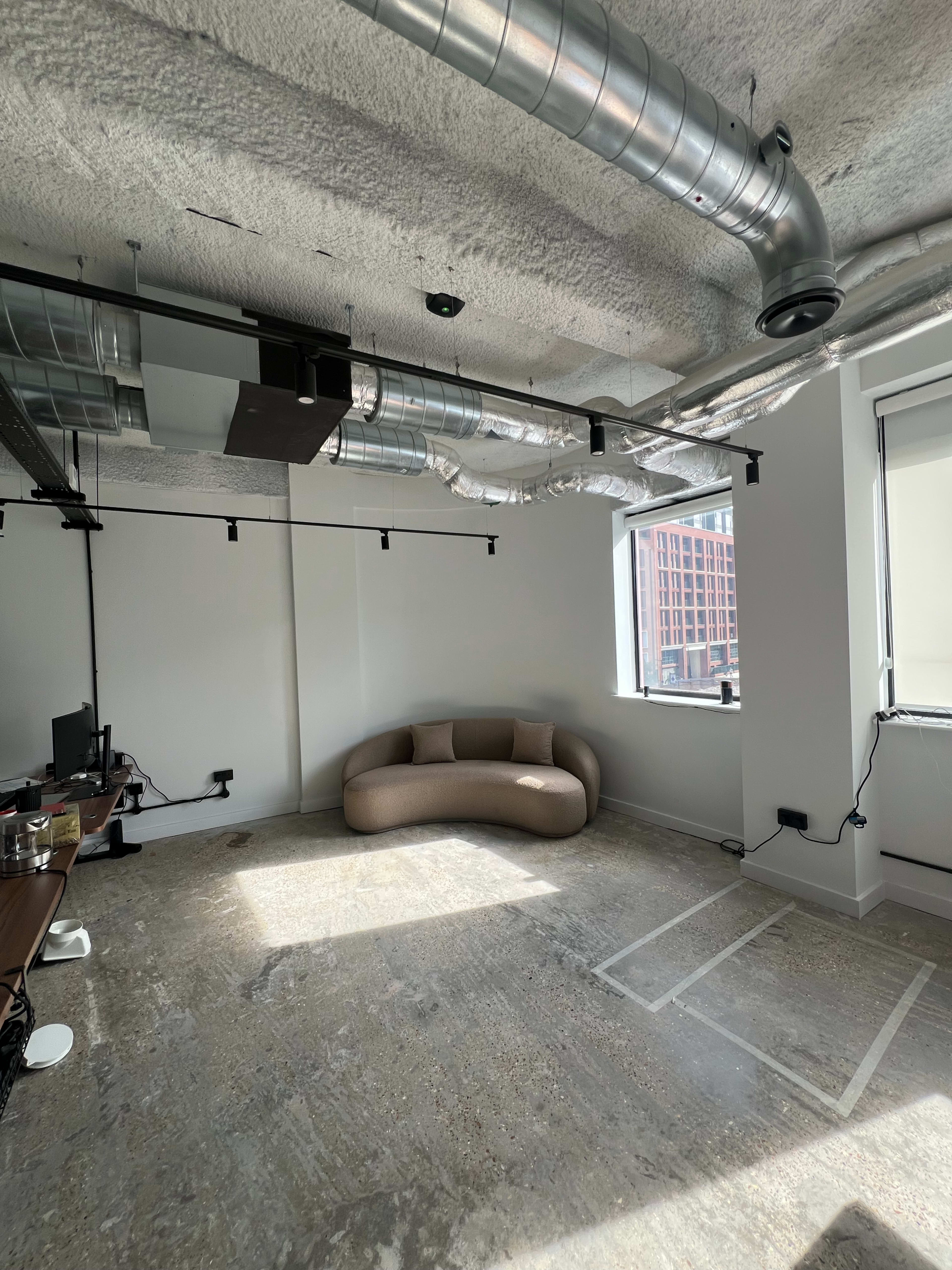The image shows an empty office space with a curved brown sofa in one corner and exposed ventilation ducts on the ceiling.