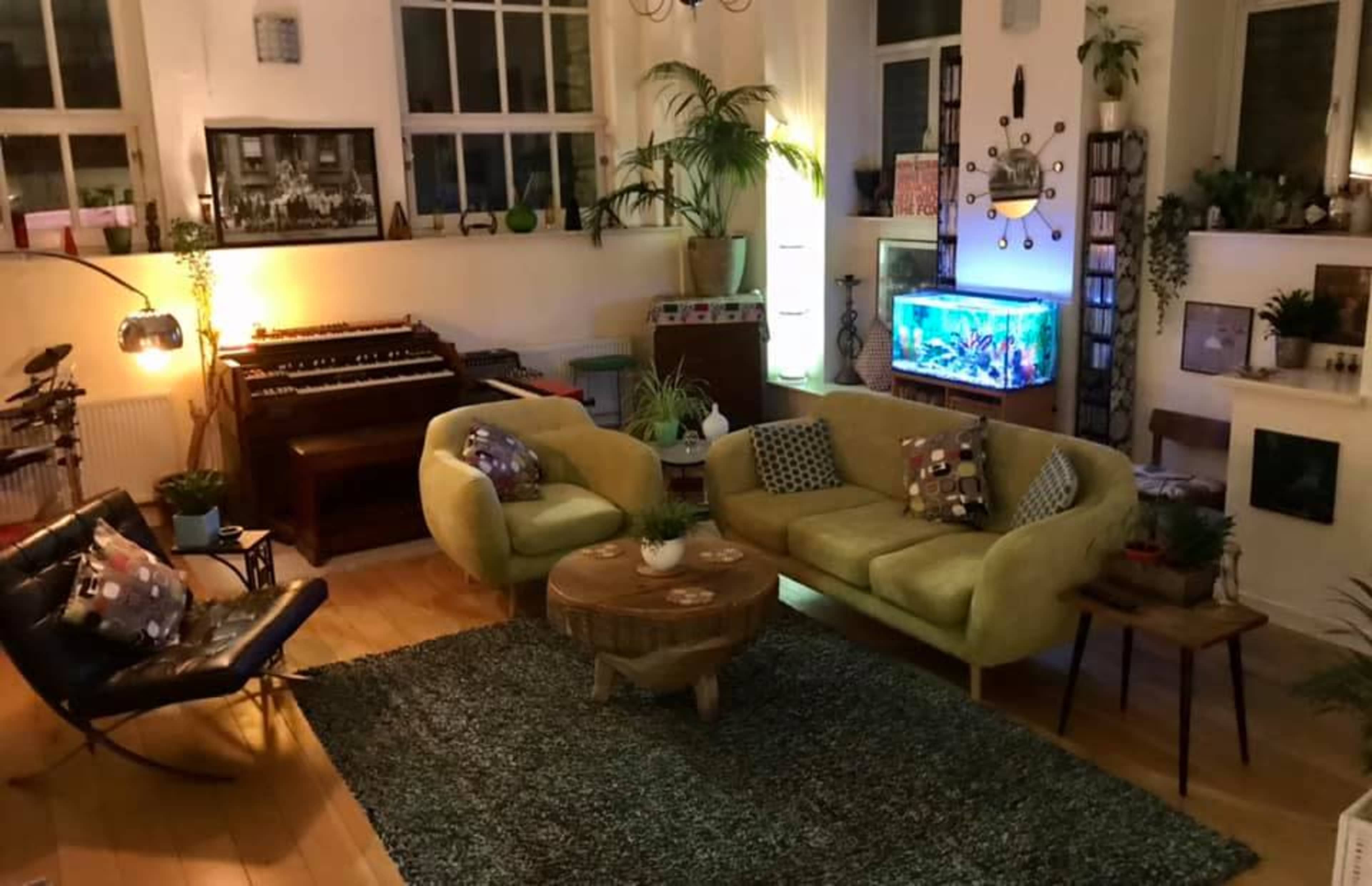 The image shows a cozy living room with a yellow sofa, a round wooden coffee table, an aquarium, and plants, set against a backdrop of large windows and various decorative elements.