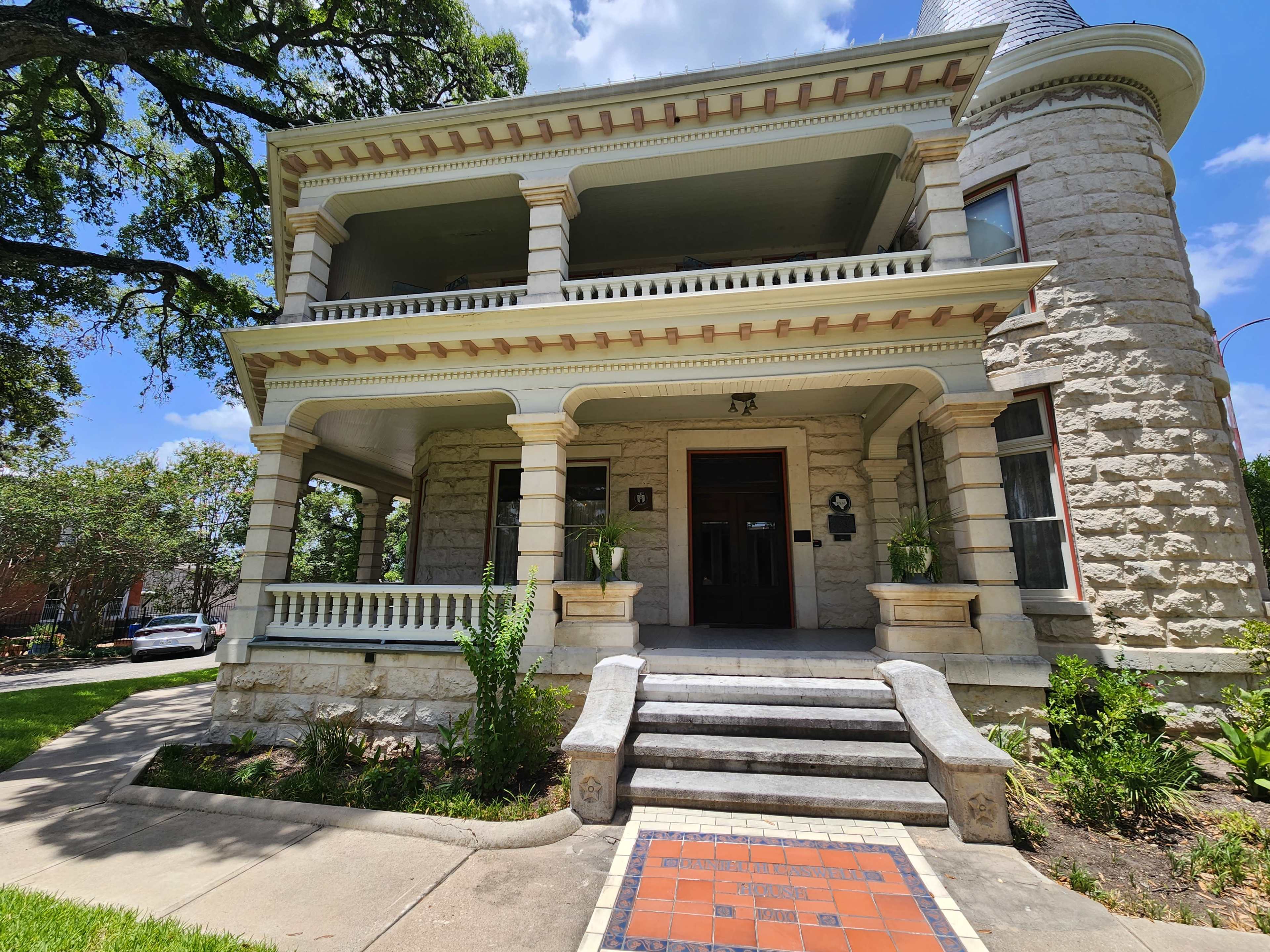The image shows a historic stone building with two levels, featuring a grand entrance and decorative columns, surrounded by greenery and a pathway leading to the front steps.