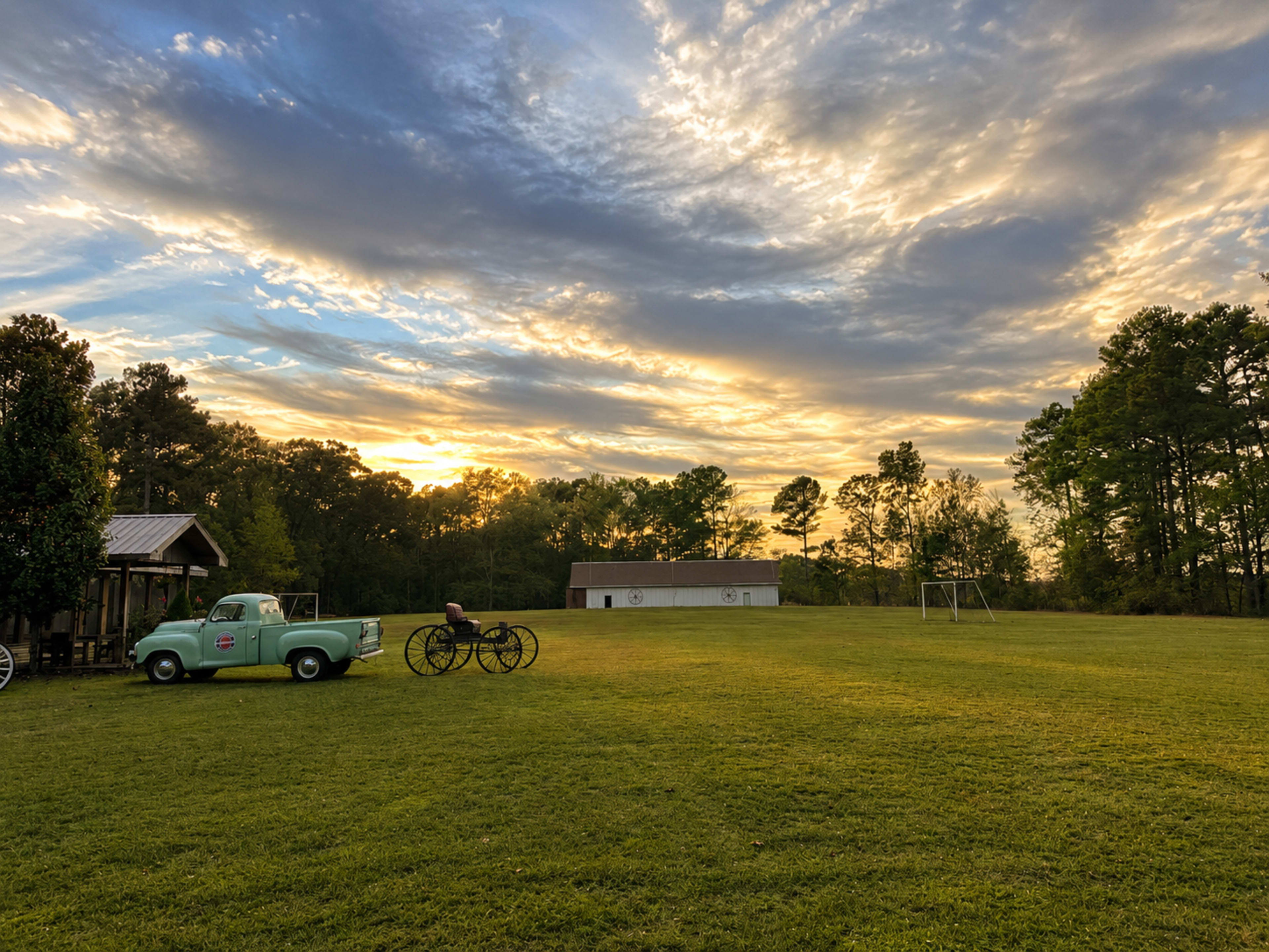 A blue vintage truck and a horse-drawn carriage are positioned on a grassy field under a colorful sunset sky with scattered clouds.