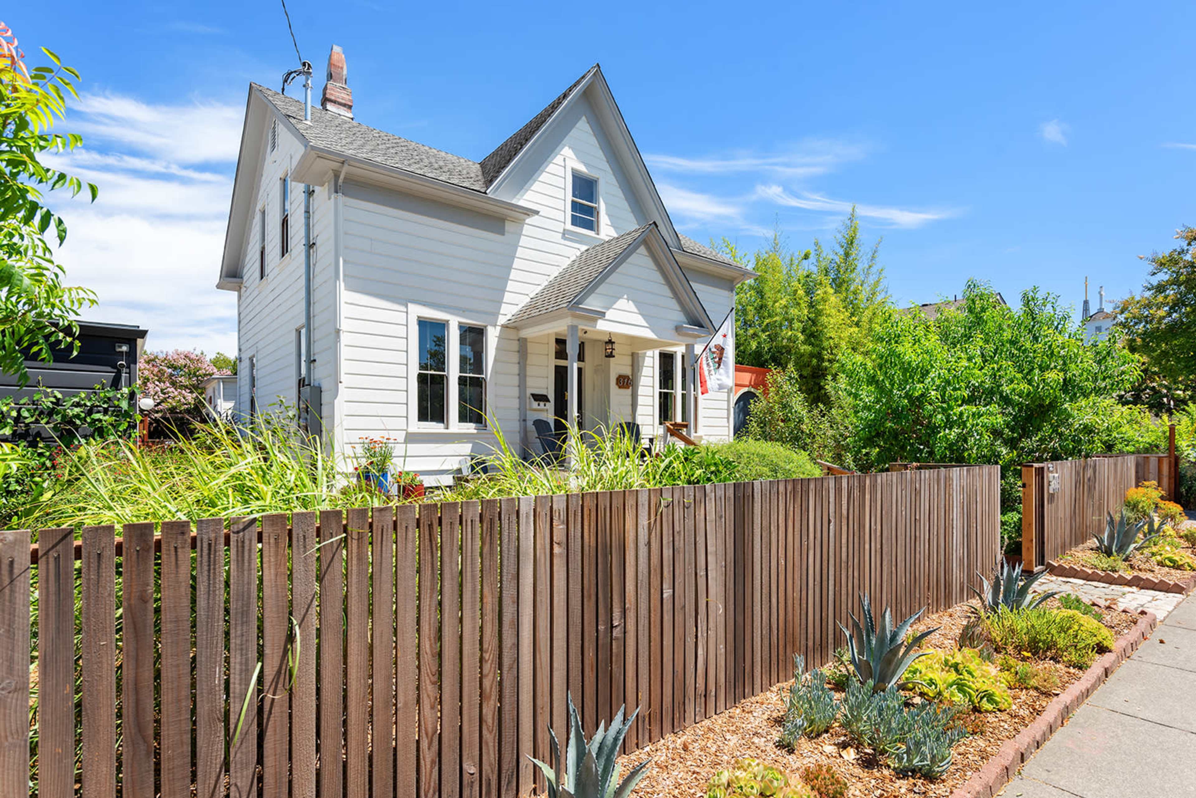 A white two-story house with a front porch is surrounded by a wooden fence and landscaped plants.