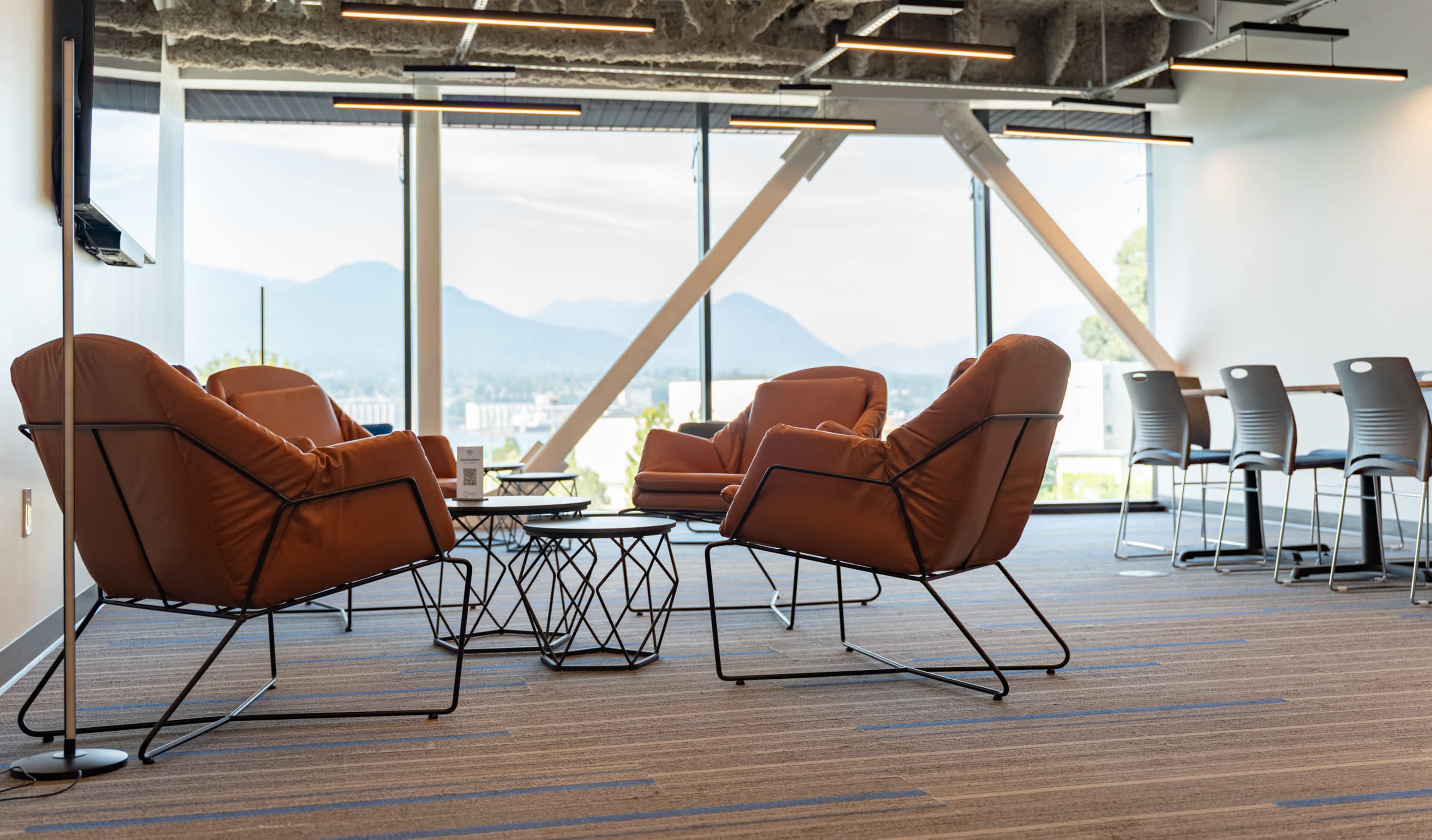 The image features a modern lounge area with four brown armchairs arranged around a small table, set against large windows that overlook a mountainous landscape.
