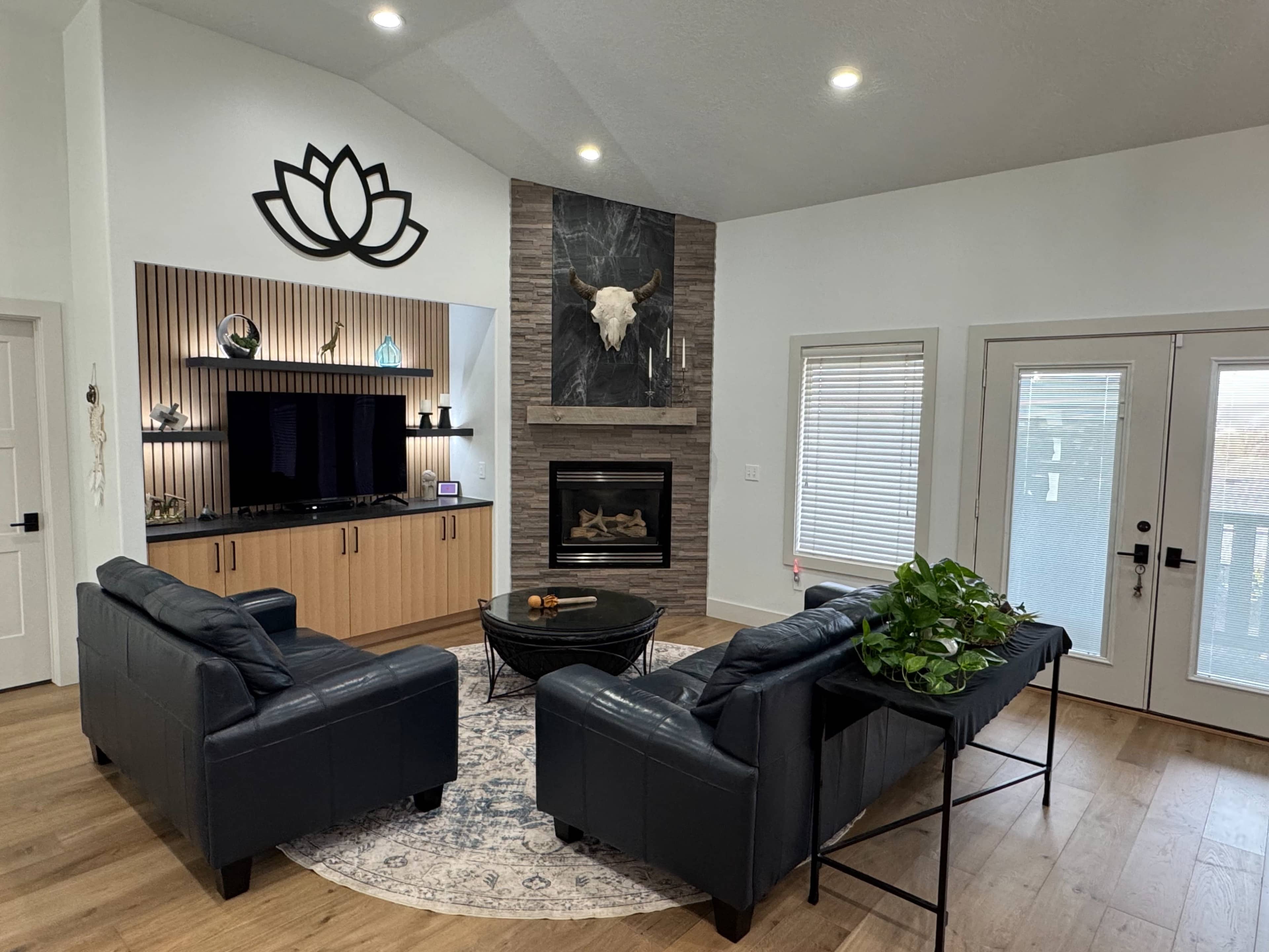 A modern living room features a black leather seating arrangement, a large television set against a wooden wall, and a fireplace with a decorative bull skull above it.