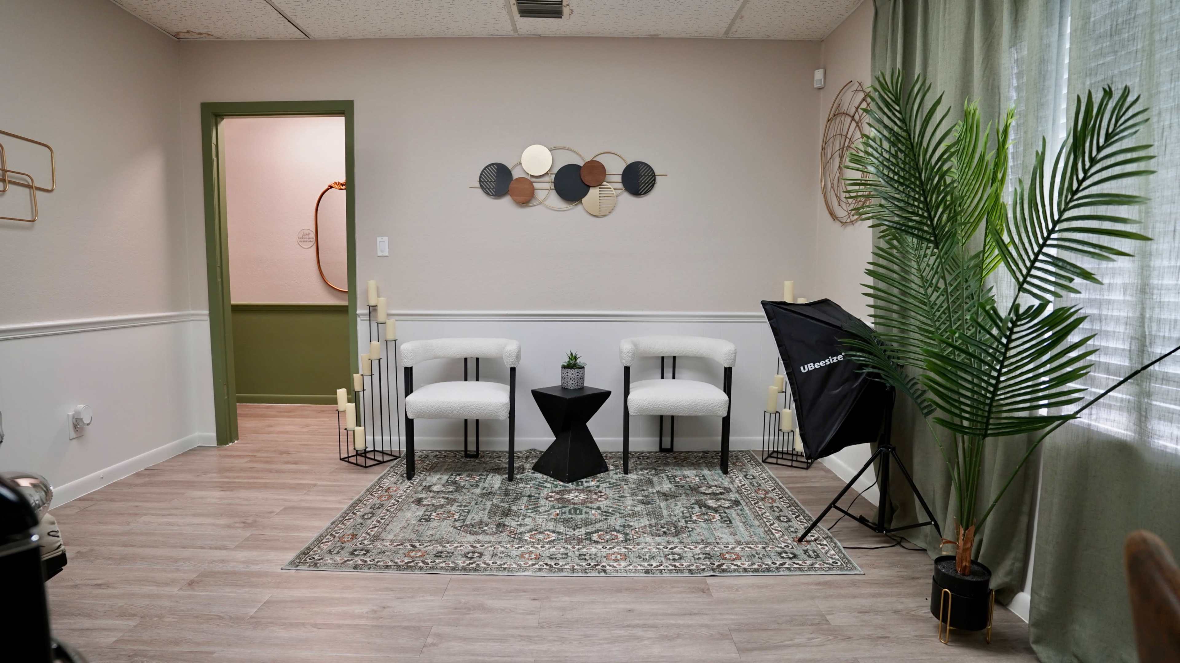 The image shows a modern room with two white chairs and a small black table on a patterned rug, accented by a plant and minimalist wall decor.
