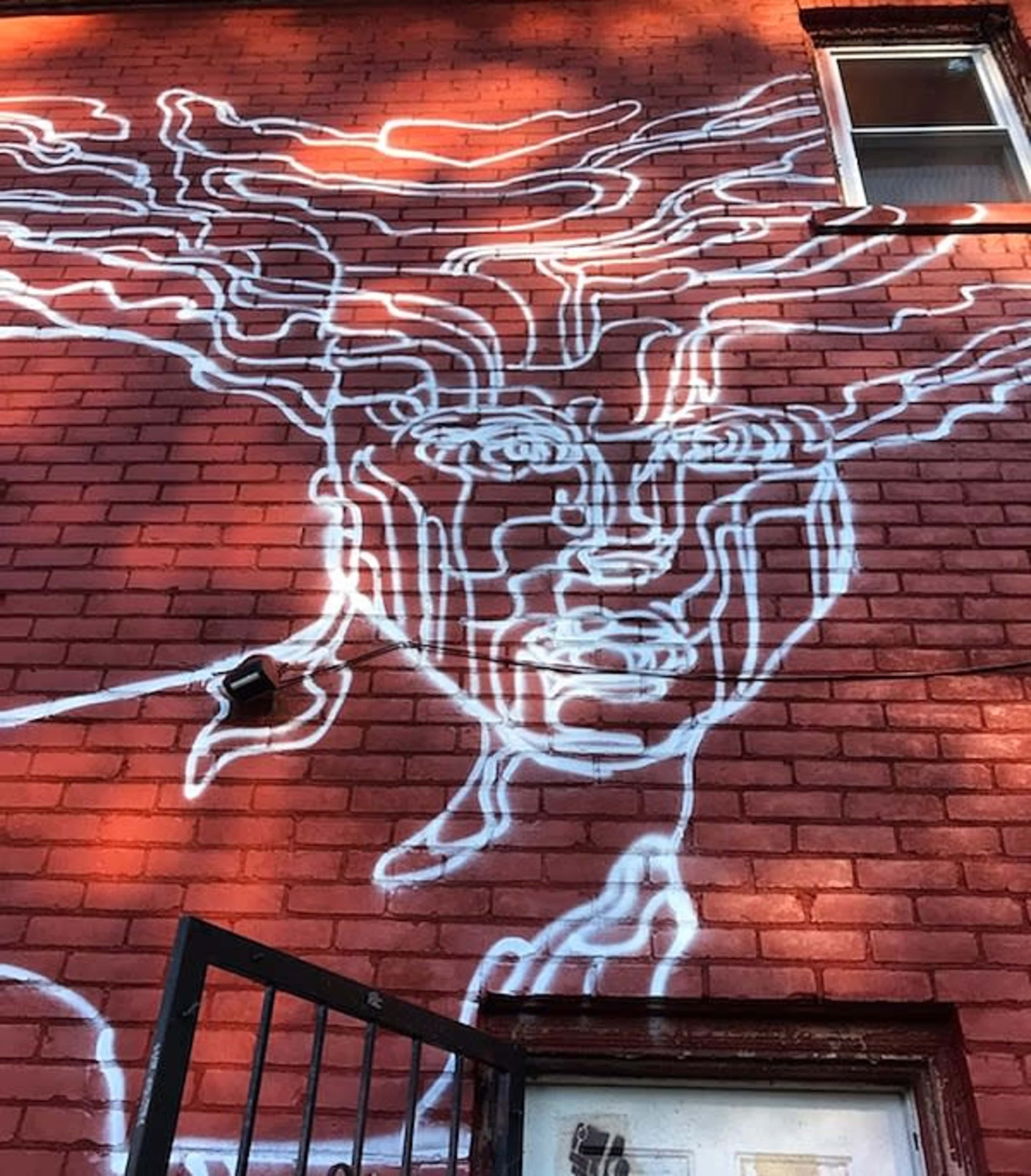 A white outlined mural depicting a face with wavy hair is painted on a red brick wall.