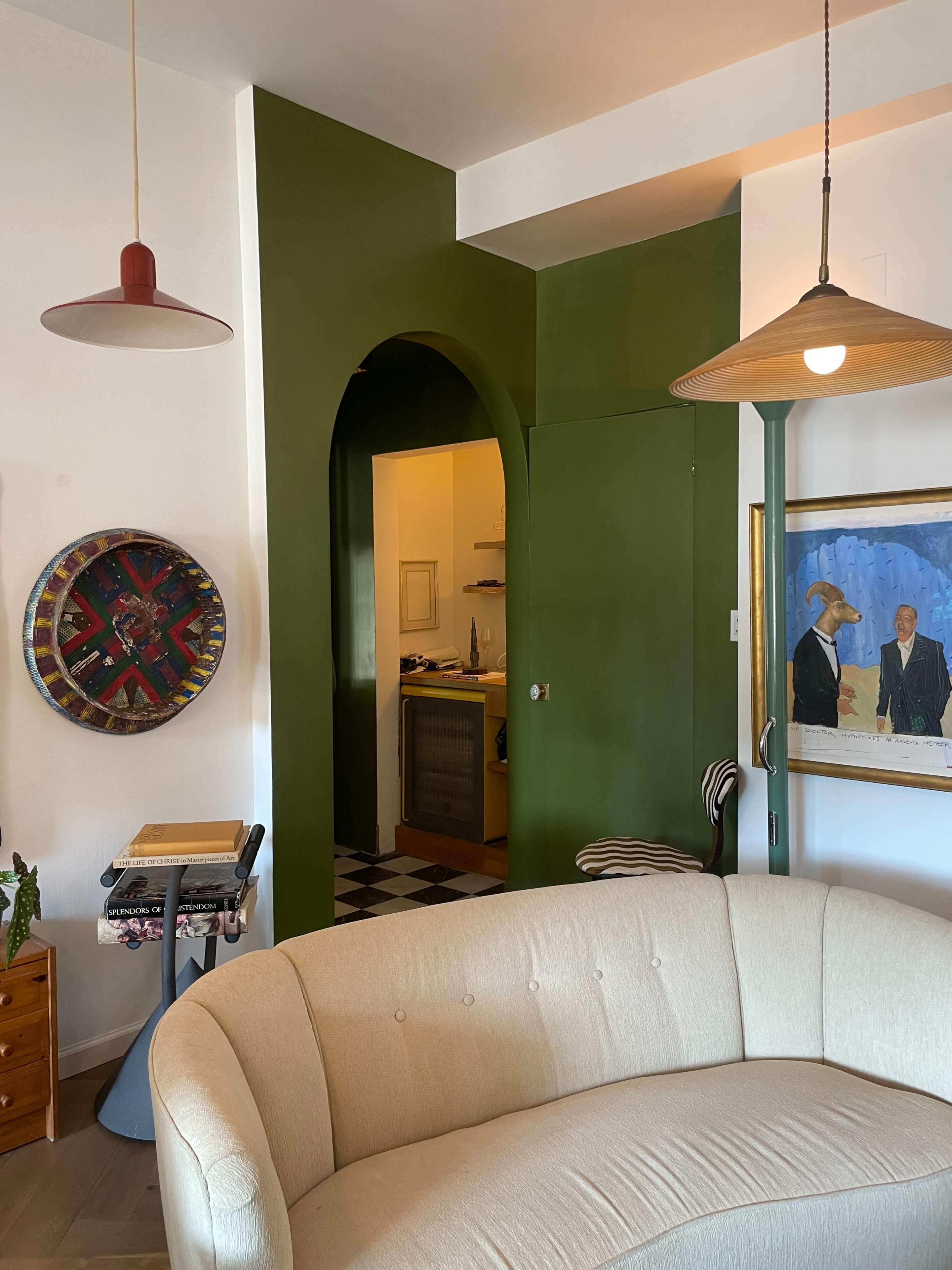 The image shows a cozy living room featuring a curved beige couch, green archway leading to a kitchen area, and framed artworks on the walls.