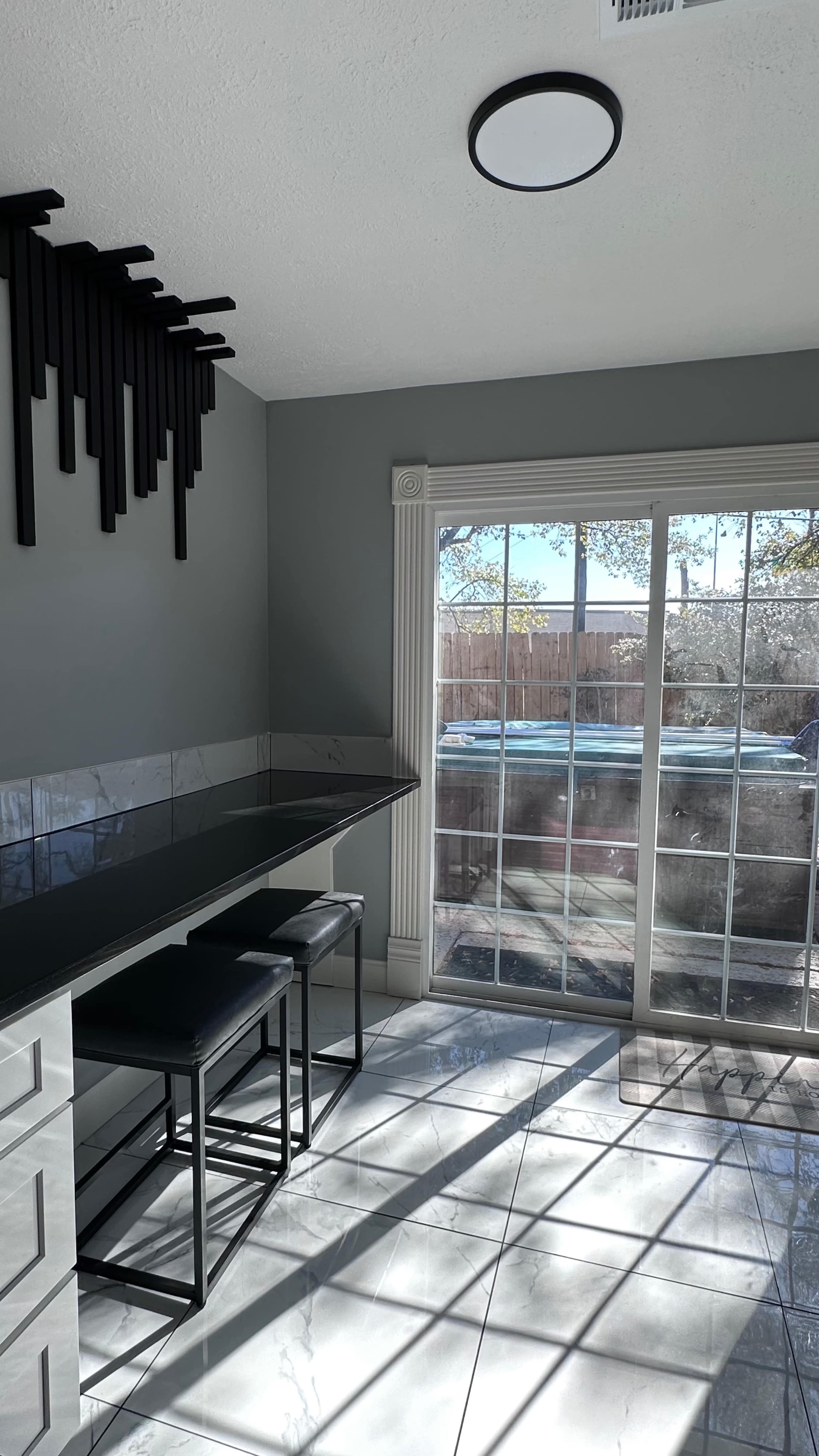 A bright room features a glass sliding door leading to an outdoor area, with a sleek black bar counter and two stools beside it.