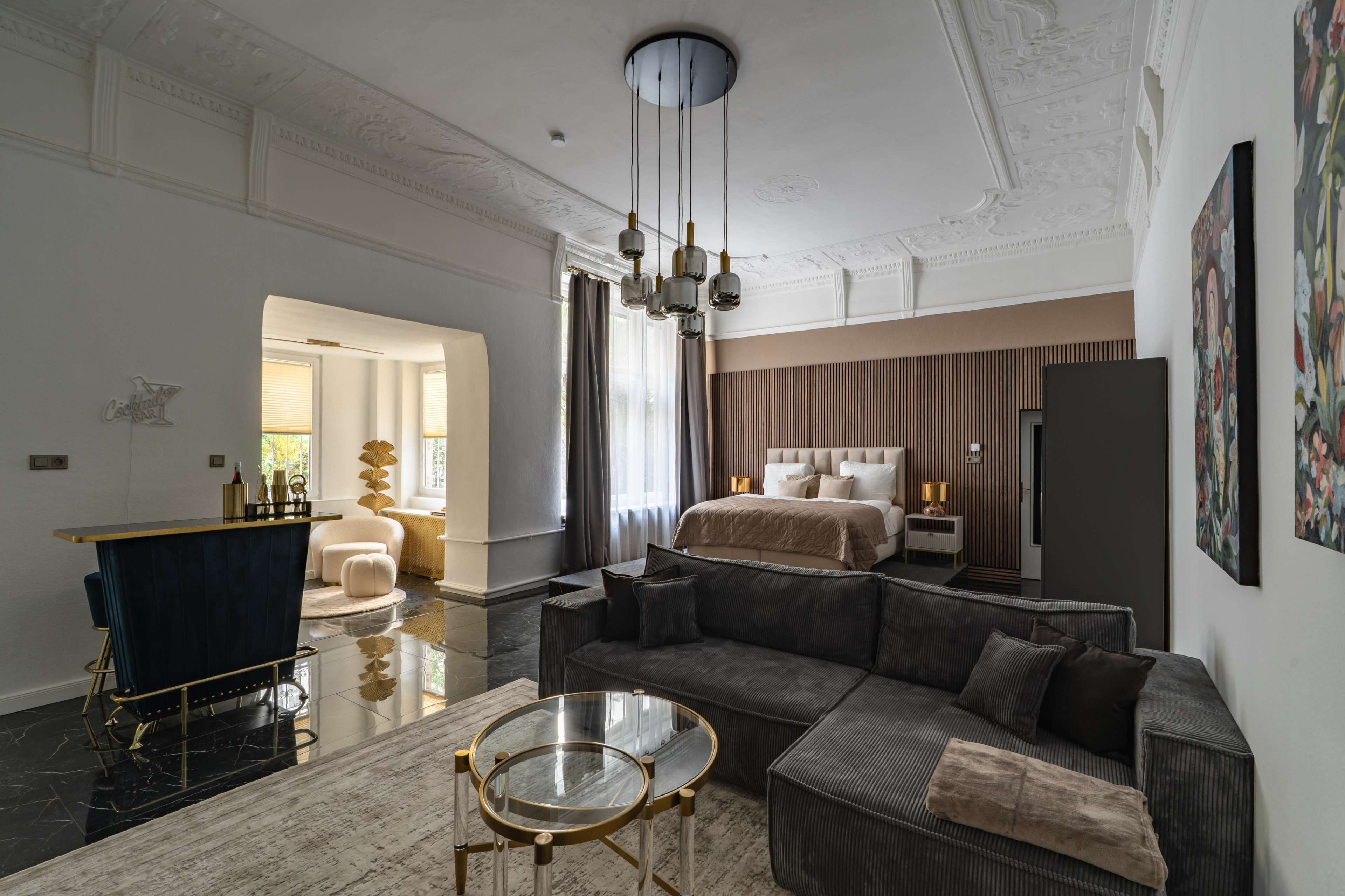 The image showcases a modern hotel suite featuring a living area with a gray sofa, a glass coffee table, and a bedroom area with a large bed and decorative wall elements.