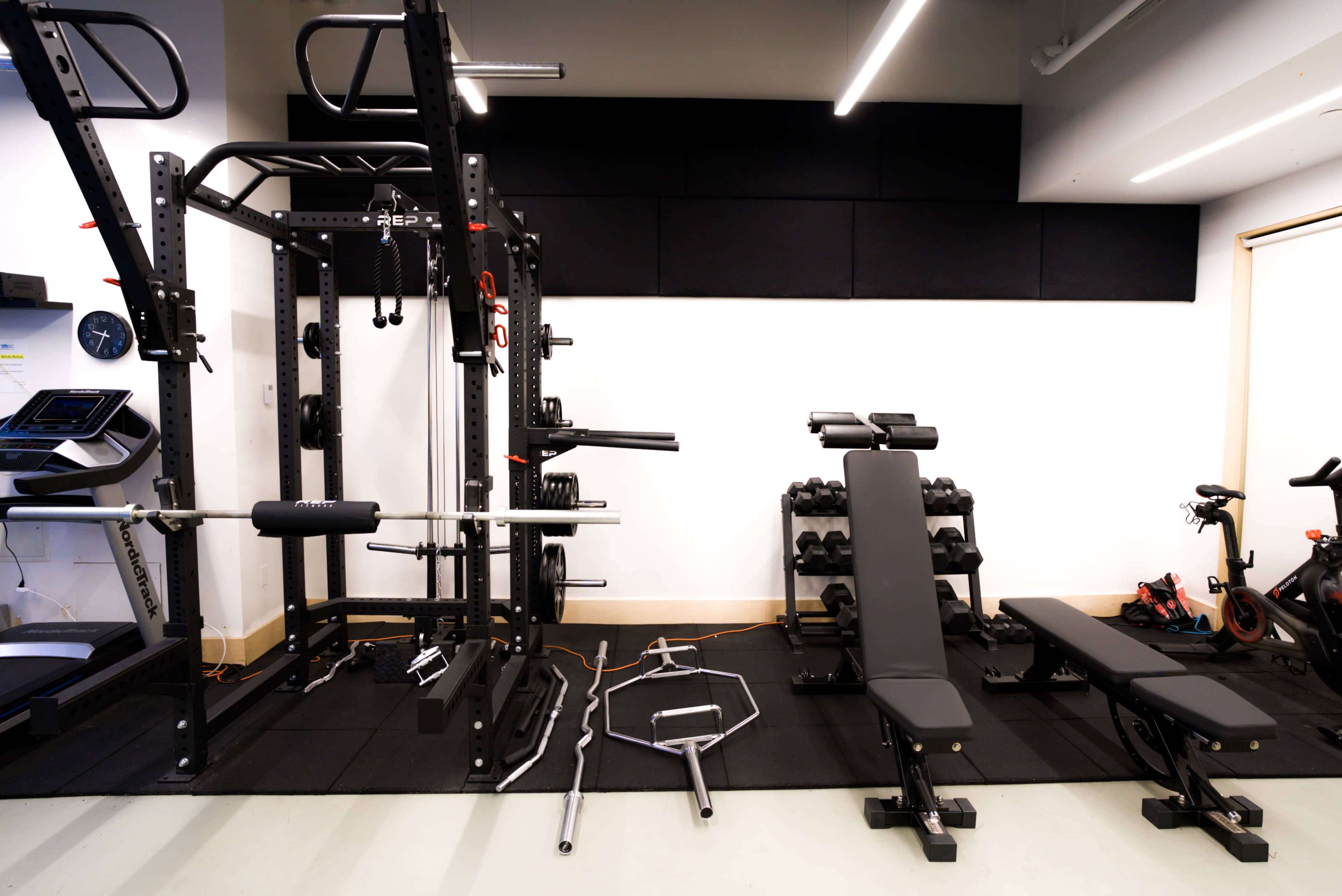 A gym space featuring a squat rack, free weights, two exercise benches, and various gym equipment arranged on a rubber mat.