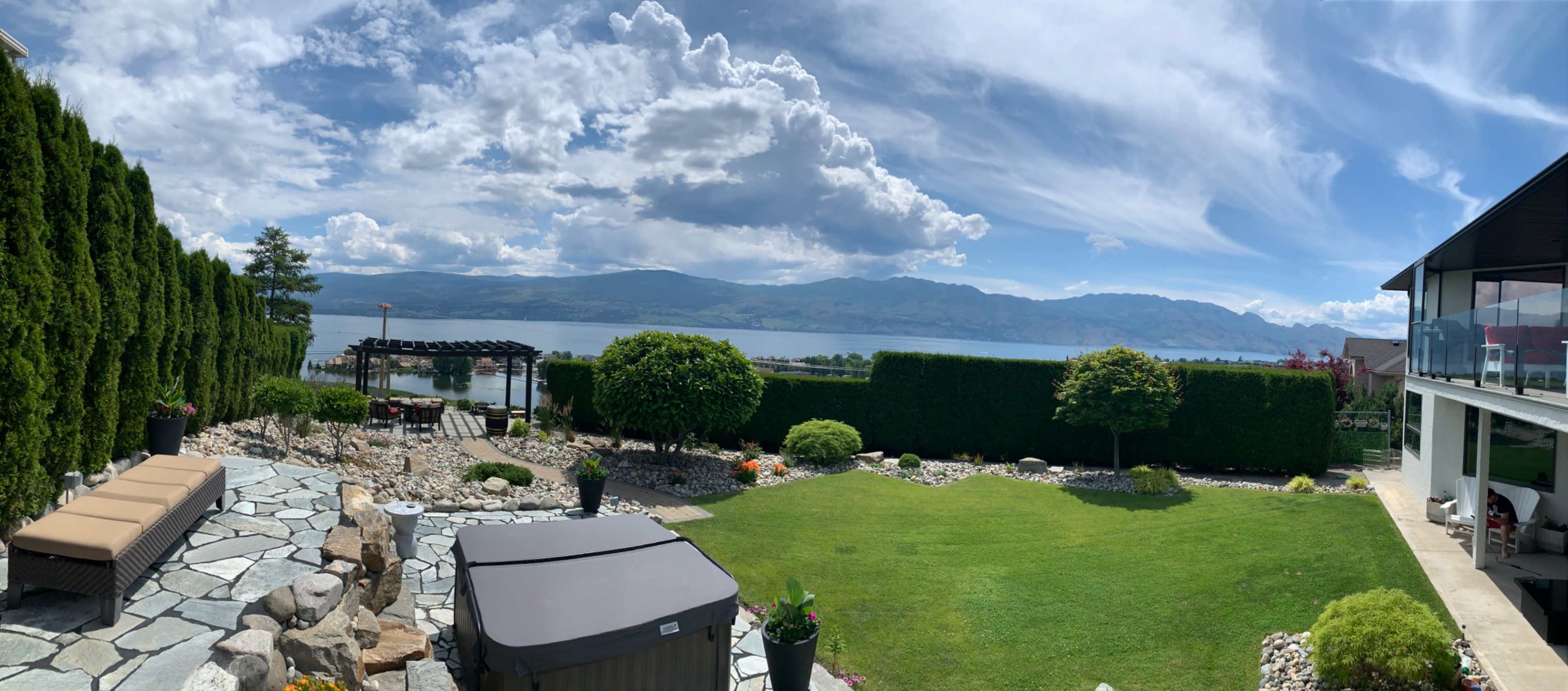 A landscaped backyard with a seating area, a hot tub, and a view of a lake surrounded by mountains under a partly cloudy sky.
