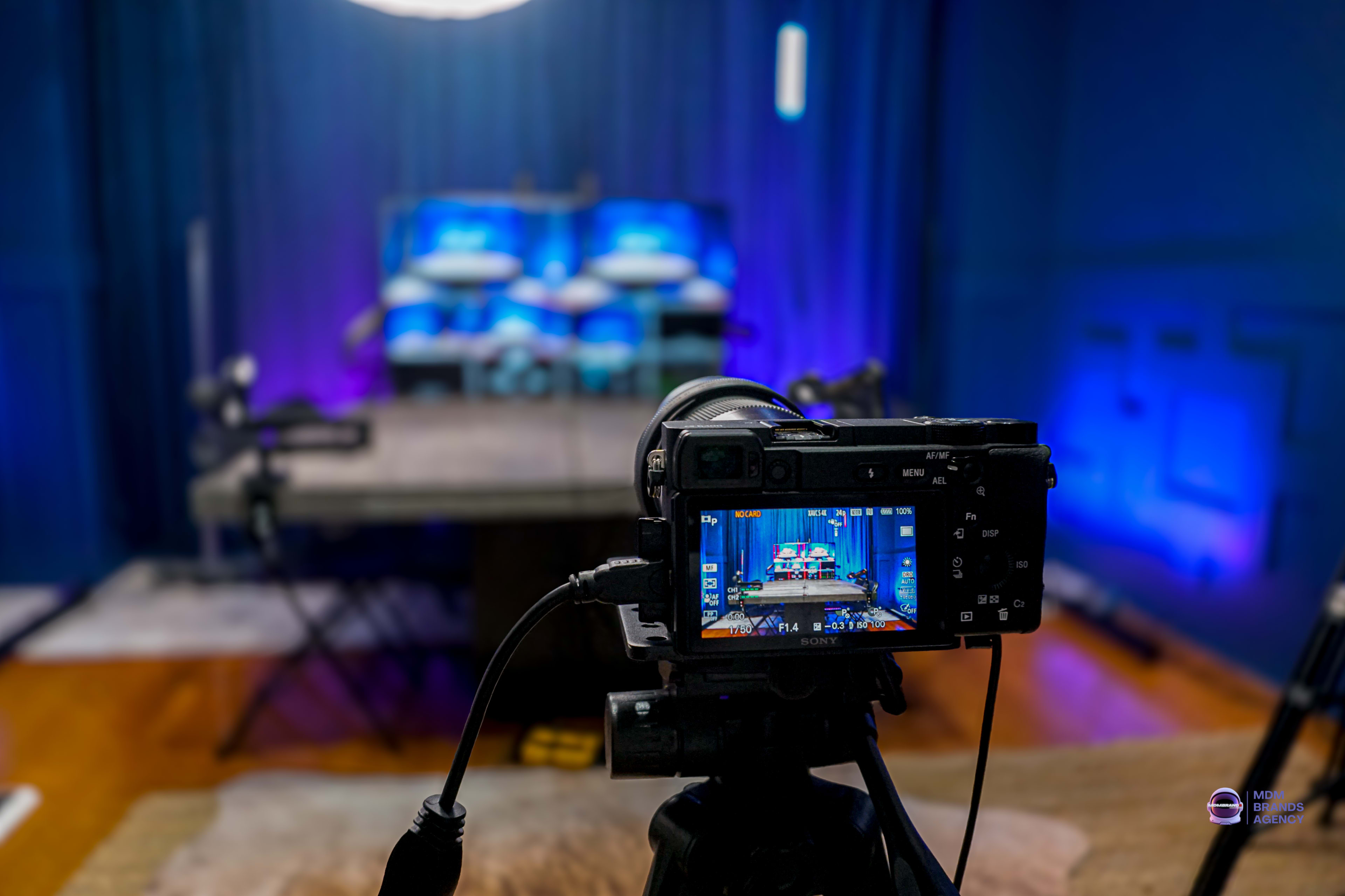 A camera is positioned on a tripod facing a stage with multiple screens illuminated by blue lighting in a room with dark walls.