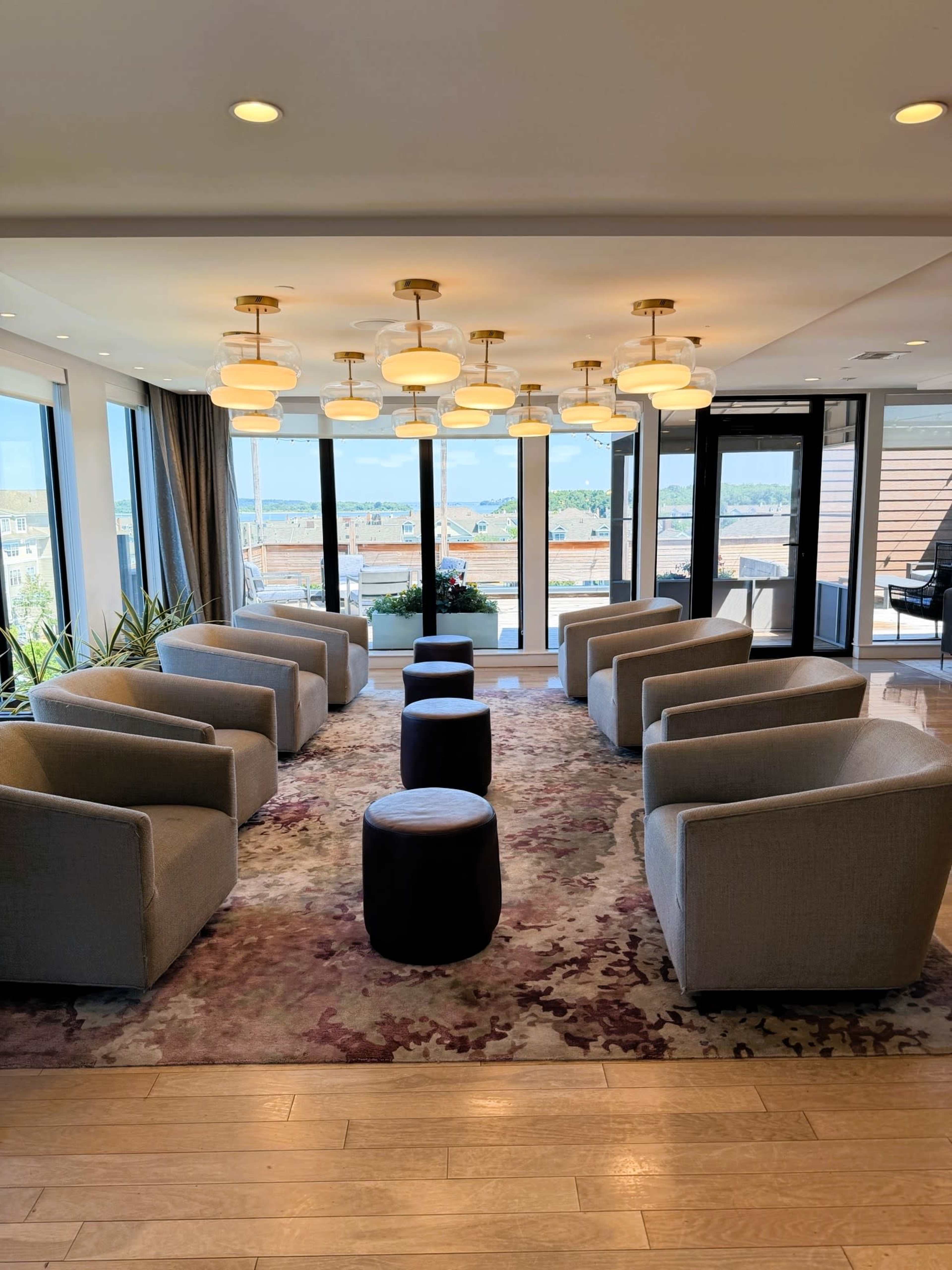 The image shows a modern lounge area with several gray armchairs, round stools, and large windows providing a view of the outside.