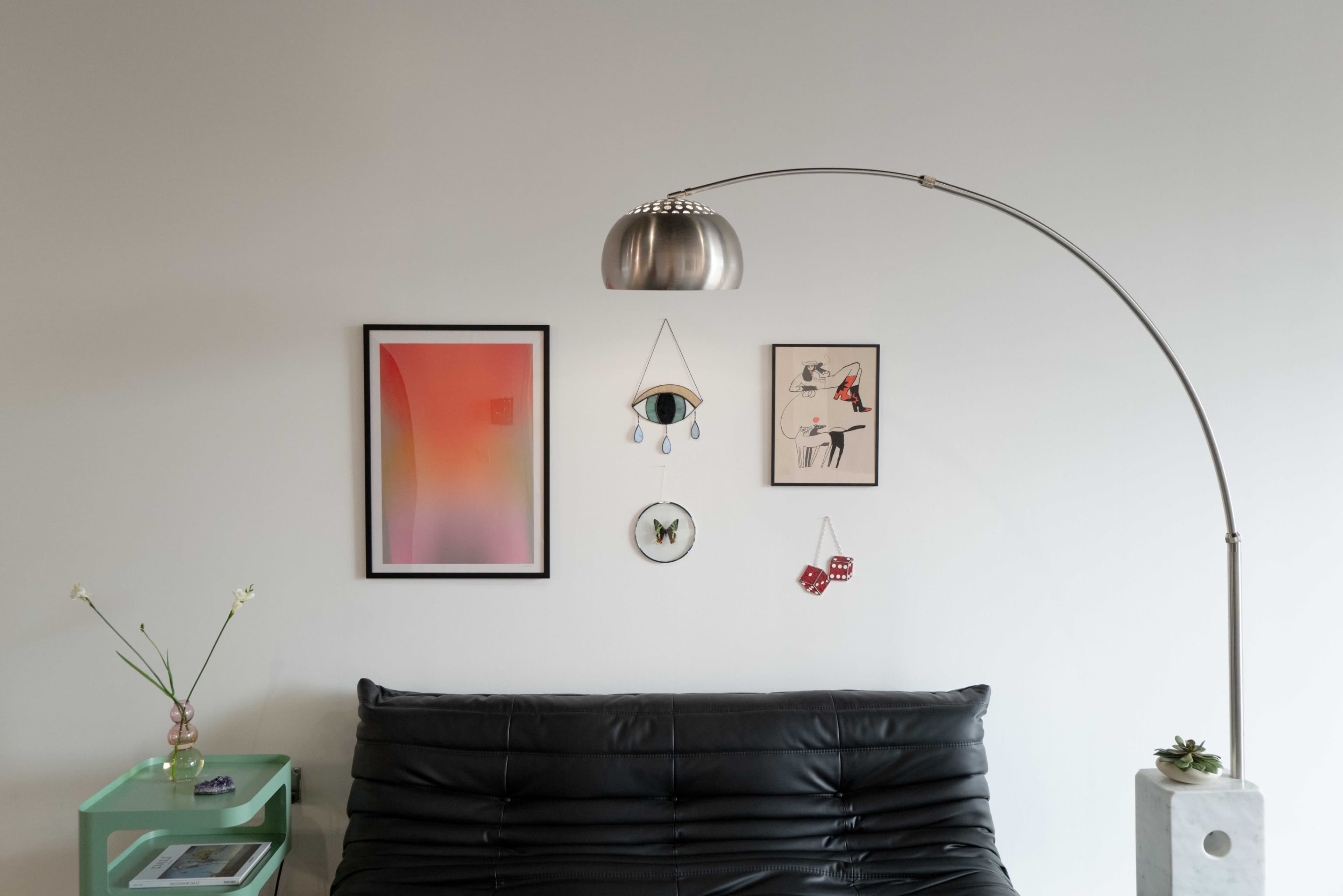 The scene features a black leather sofa underneath a sleek arc lamp, with various framed artworks and decorative items arranged on the white wall behind.