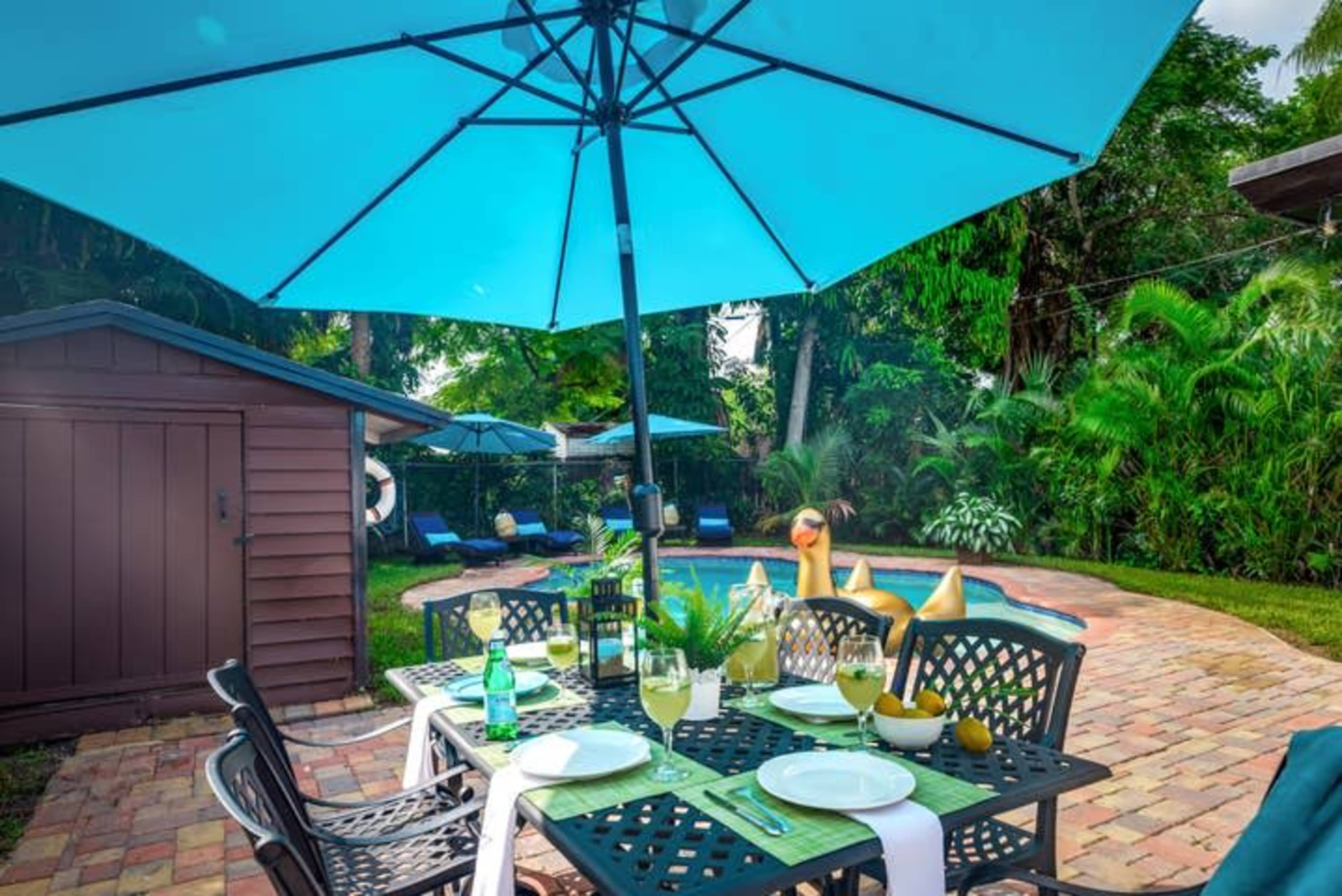 A patio table is set with drinks and snacks under a large blue umbrella next to a swimming pool surrounded by lush greenery.