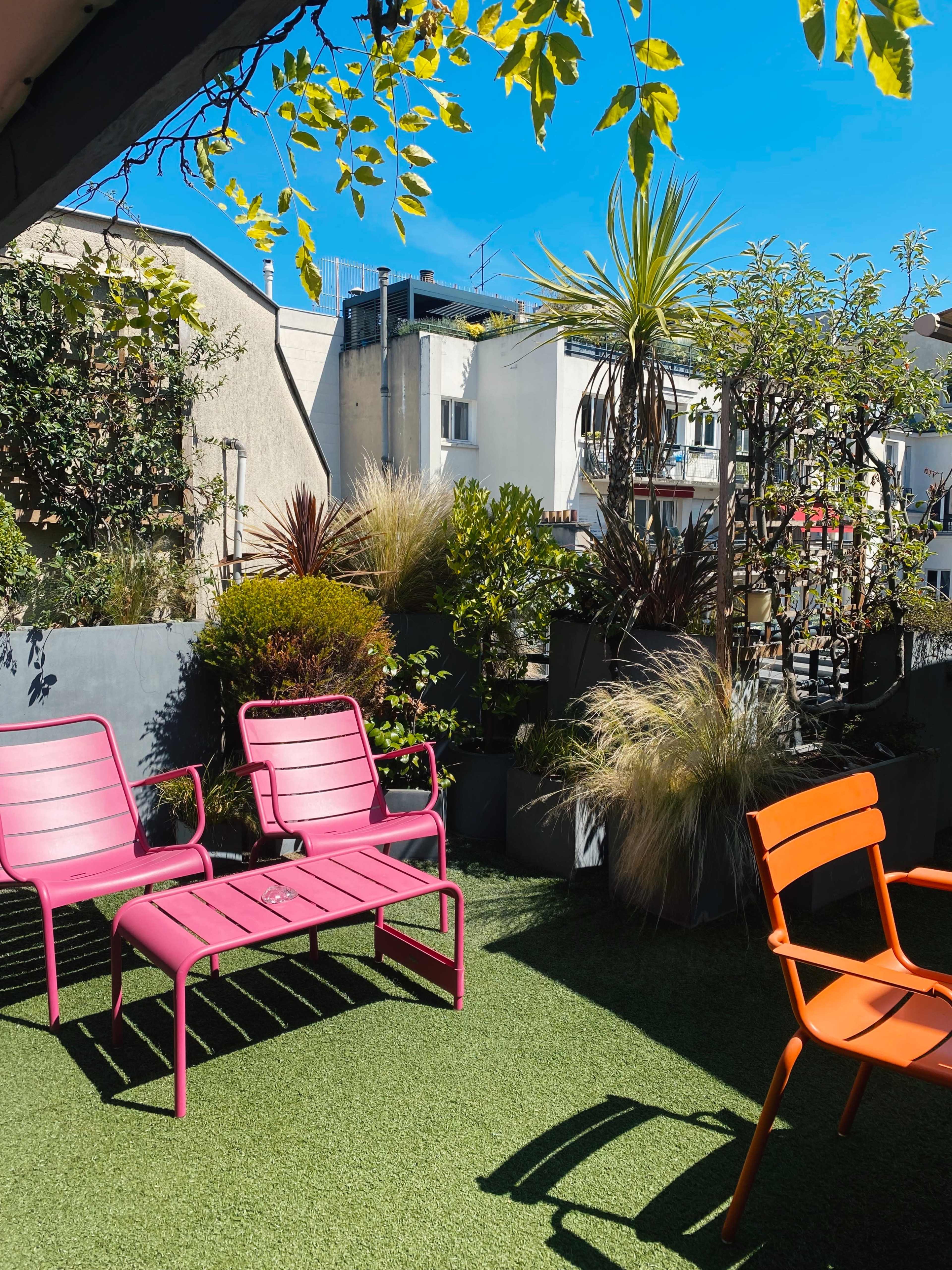A rooftop garden featuring pink and orange lounge chairs, surrounded by various plants and greenery against a backdrop of buildings and a clear blue sky.