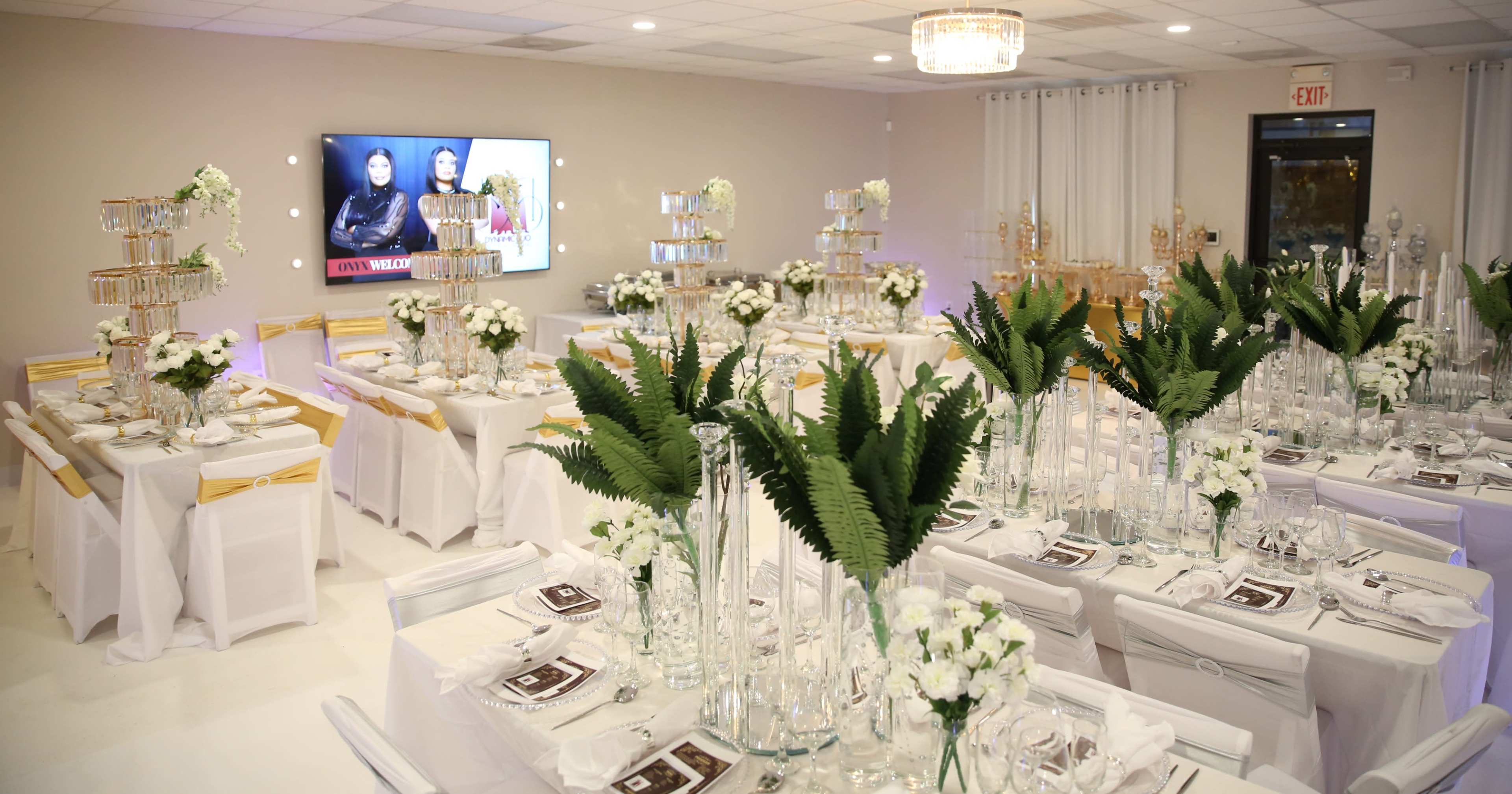 An elegantly arranged banquet hall with tables set for a formal event, featuring floral centerpieces and a large screen displaying images on the wall.