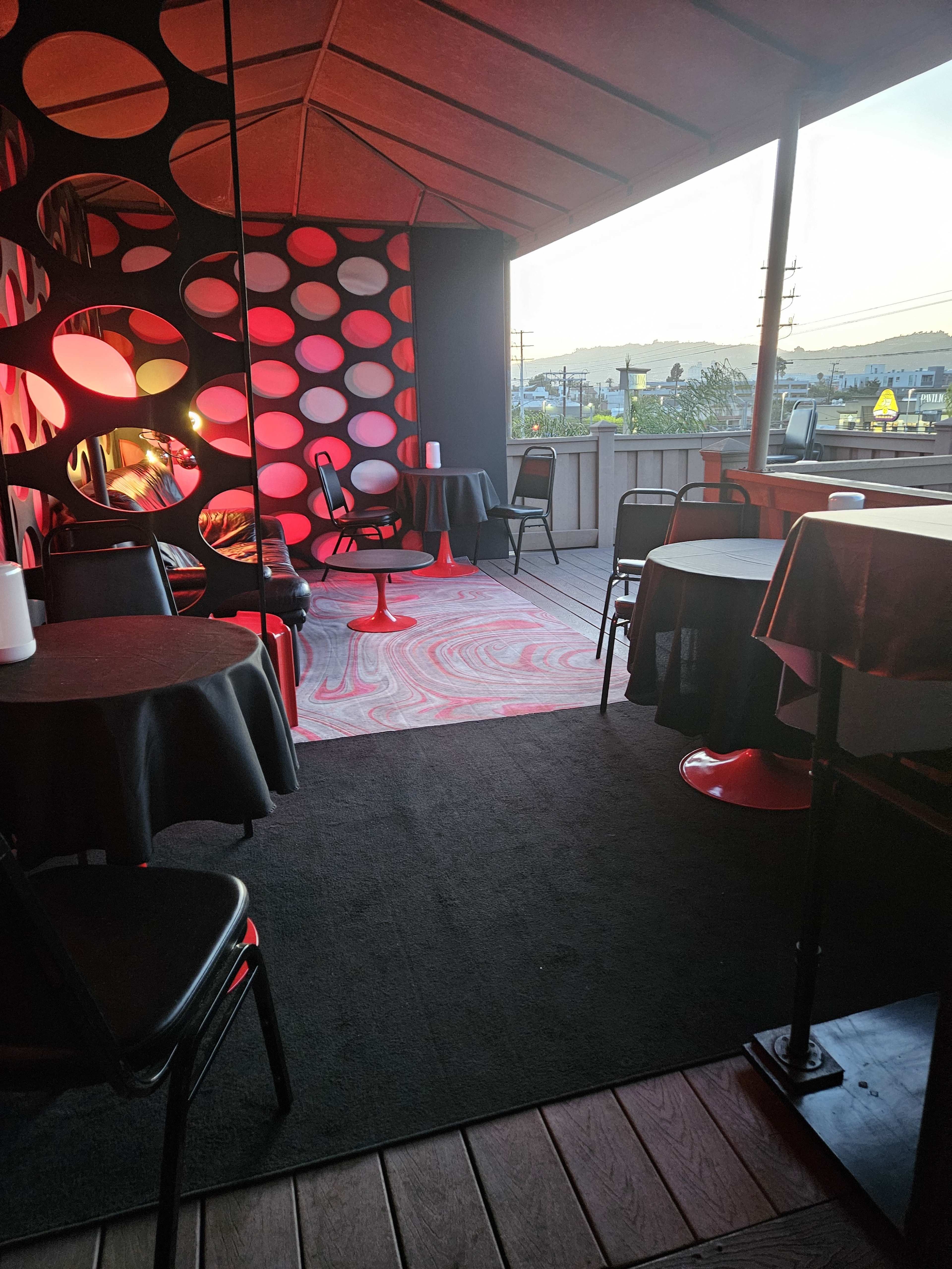 The image shows a modern outdoor seating area with circular wall decorations, red lighting, and tables set up for patrons, overlooking a distant landscape at sunset.