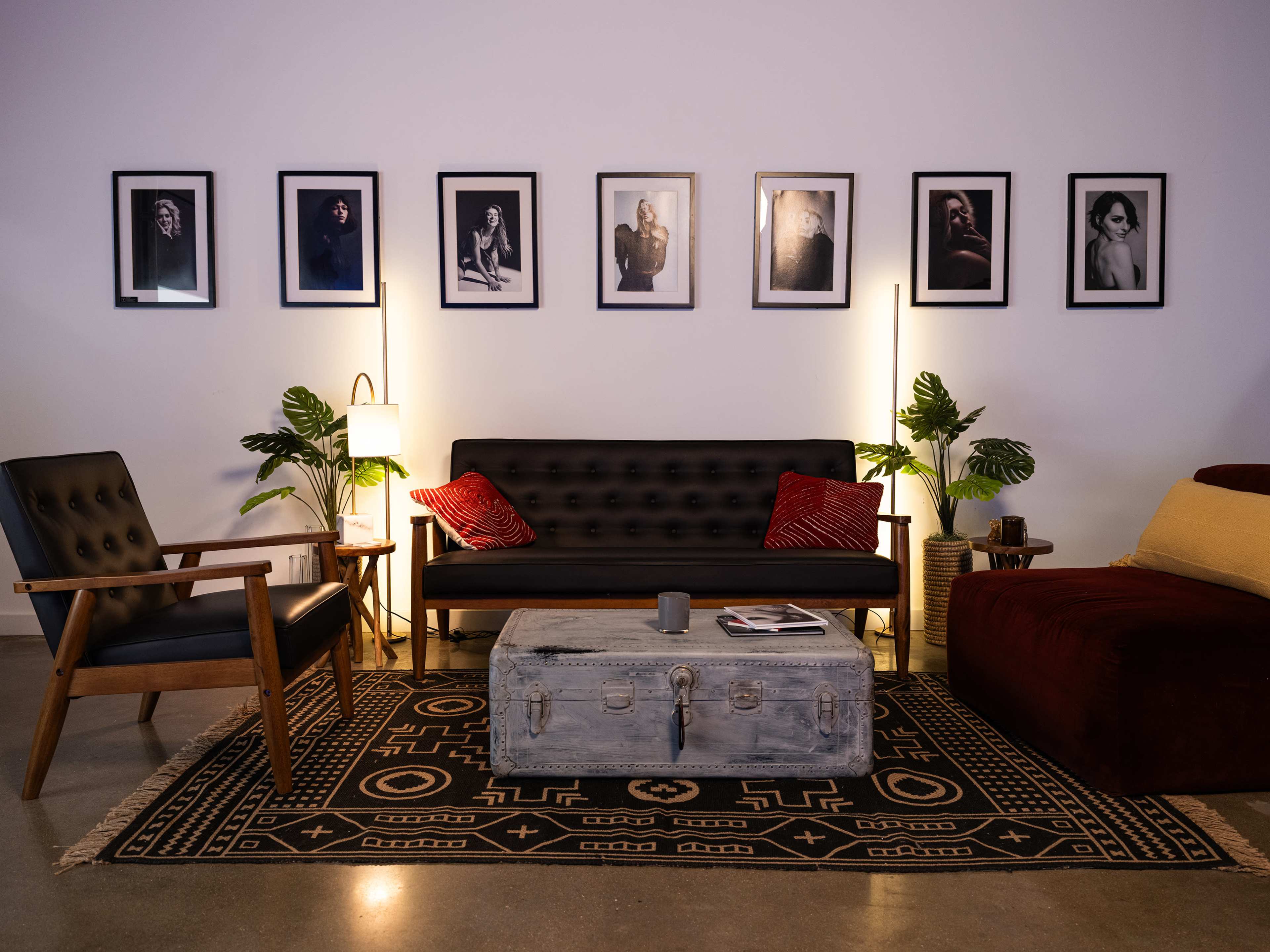 A modern living room with a black sofa, a wooden armchair, a coffee table, and framed photographs on the wall, along with plants and ambient lighting.