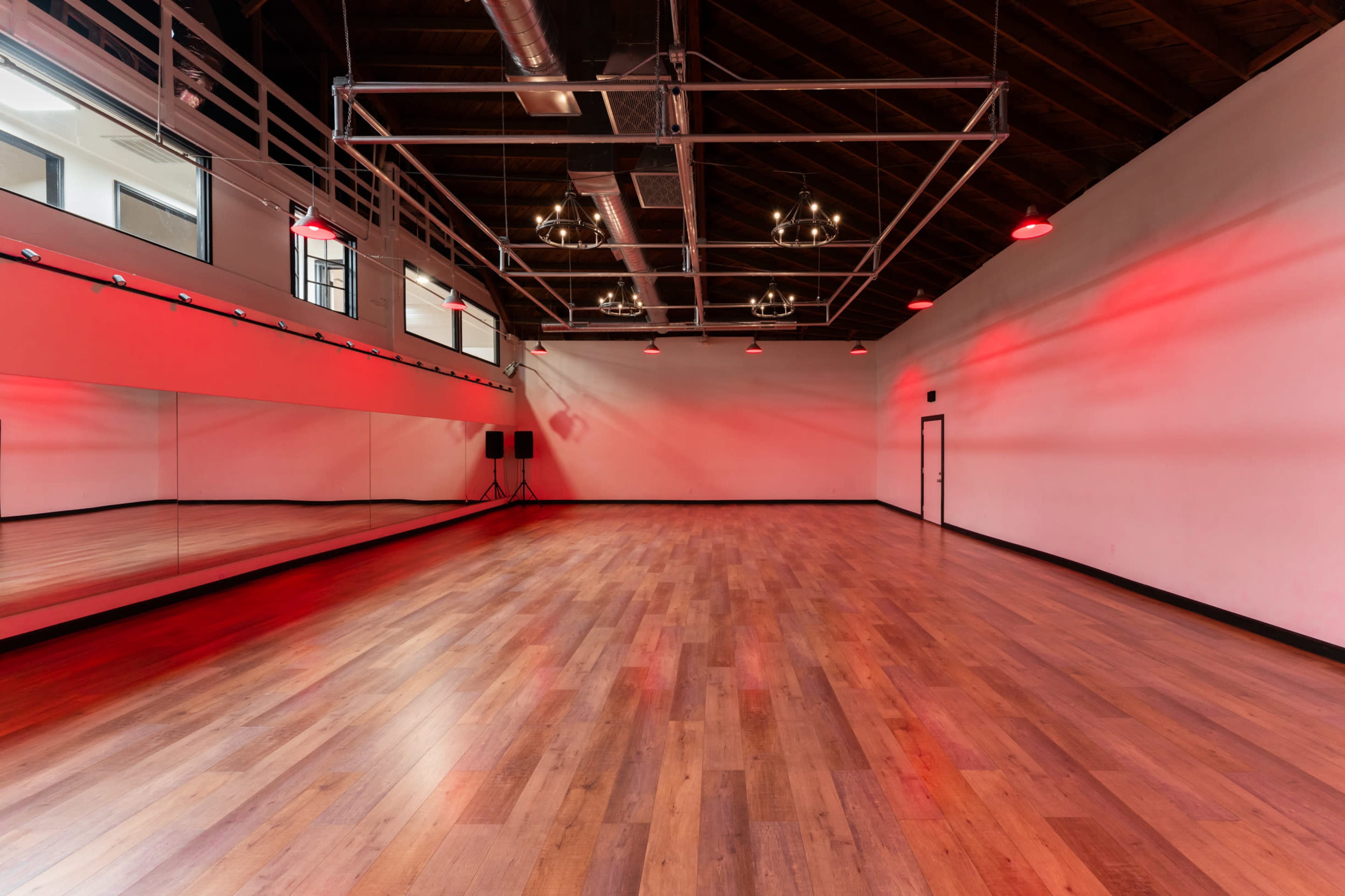 A spacious dance studio with wooden flooring, a large mirror on one wall, and red lighting fixtures along the ceiling.