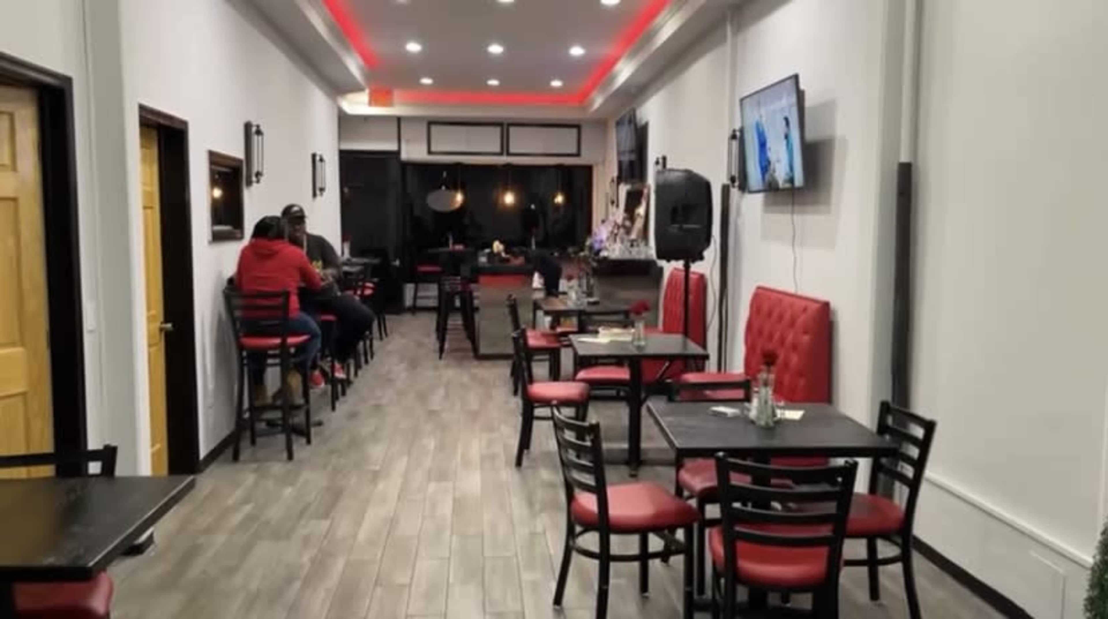 The image shows a narrow restaurant interior with several tables and chairs, and patrons sitting at the bar along one side.