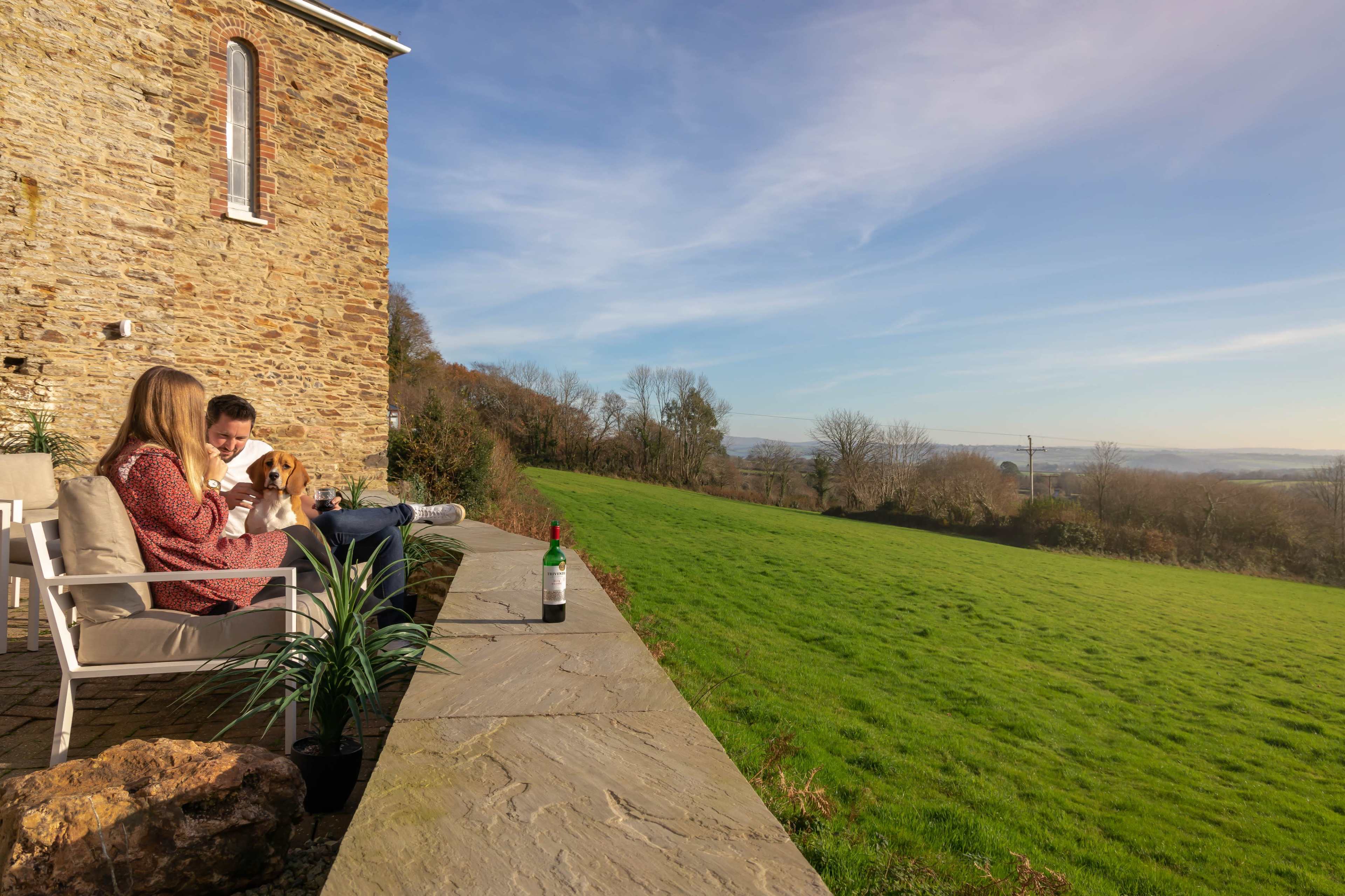 A woman and a man sit on a stone terrace with a dog and a bottle of wine, overlooking a large green field and blue sky.