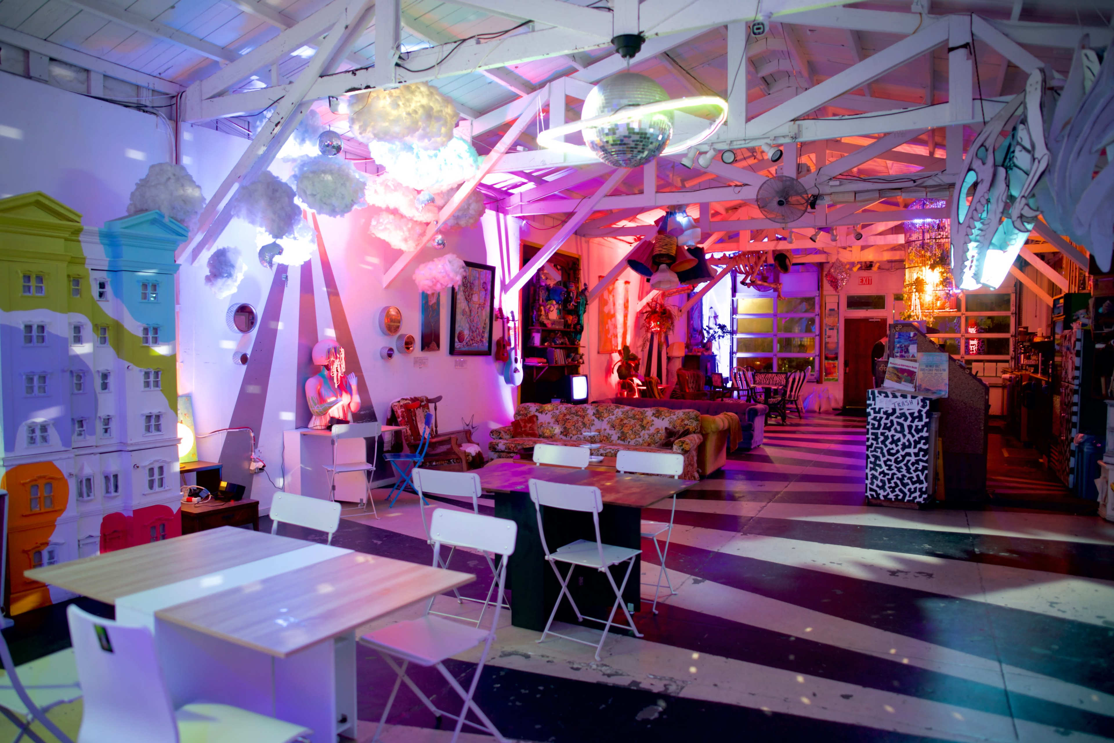 The image shows a vibrant room with colorful decor featuring white tables and chairs, artwork on the walls, and whimsical cloud-like installations hanging from the ceiling.