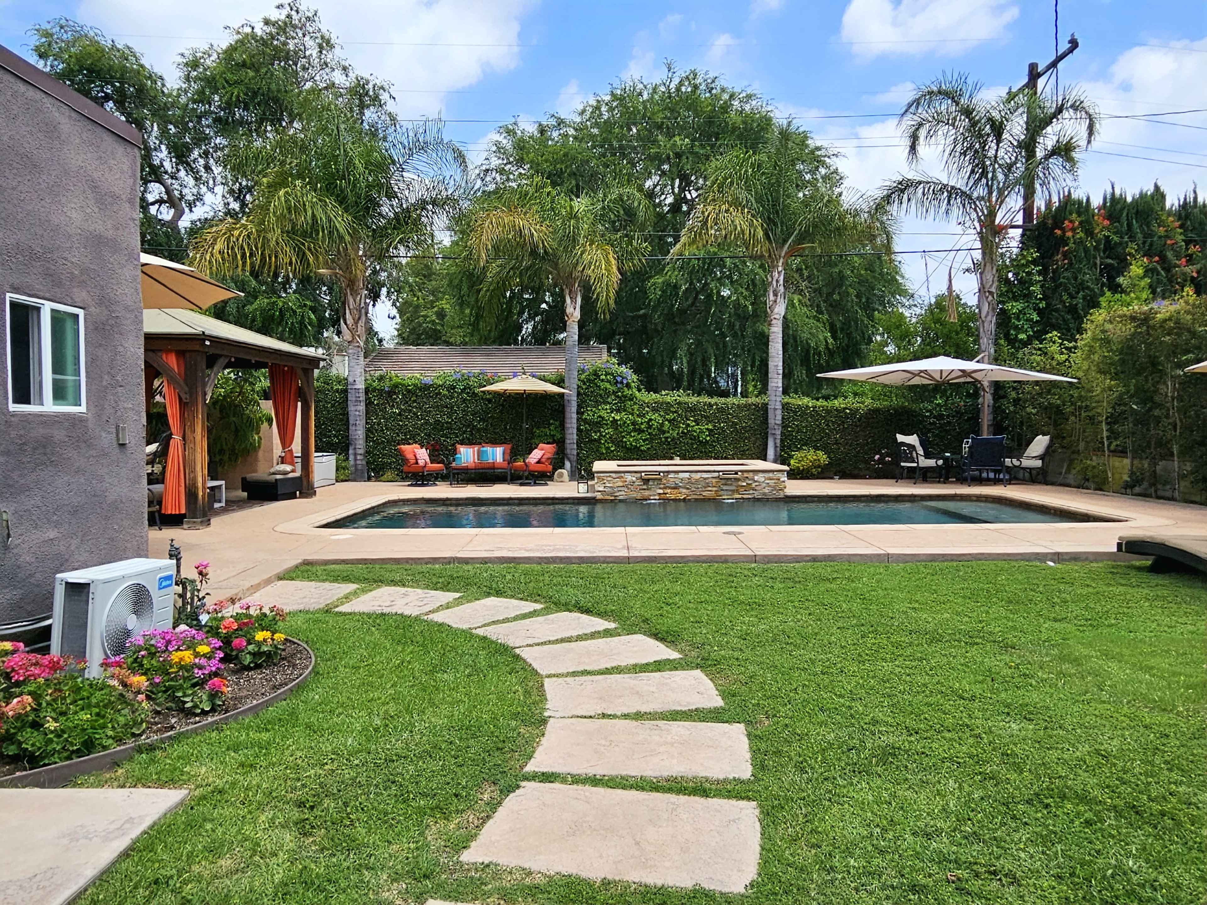 A landscaped backyard features a swimming pool surrounded by seating areas, palm trees, and colorful flower beds.