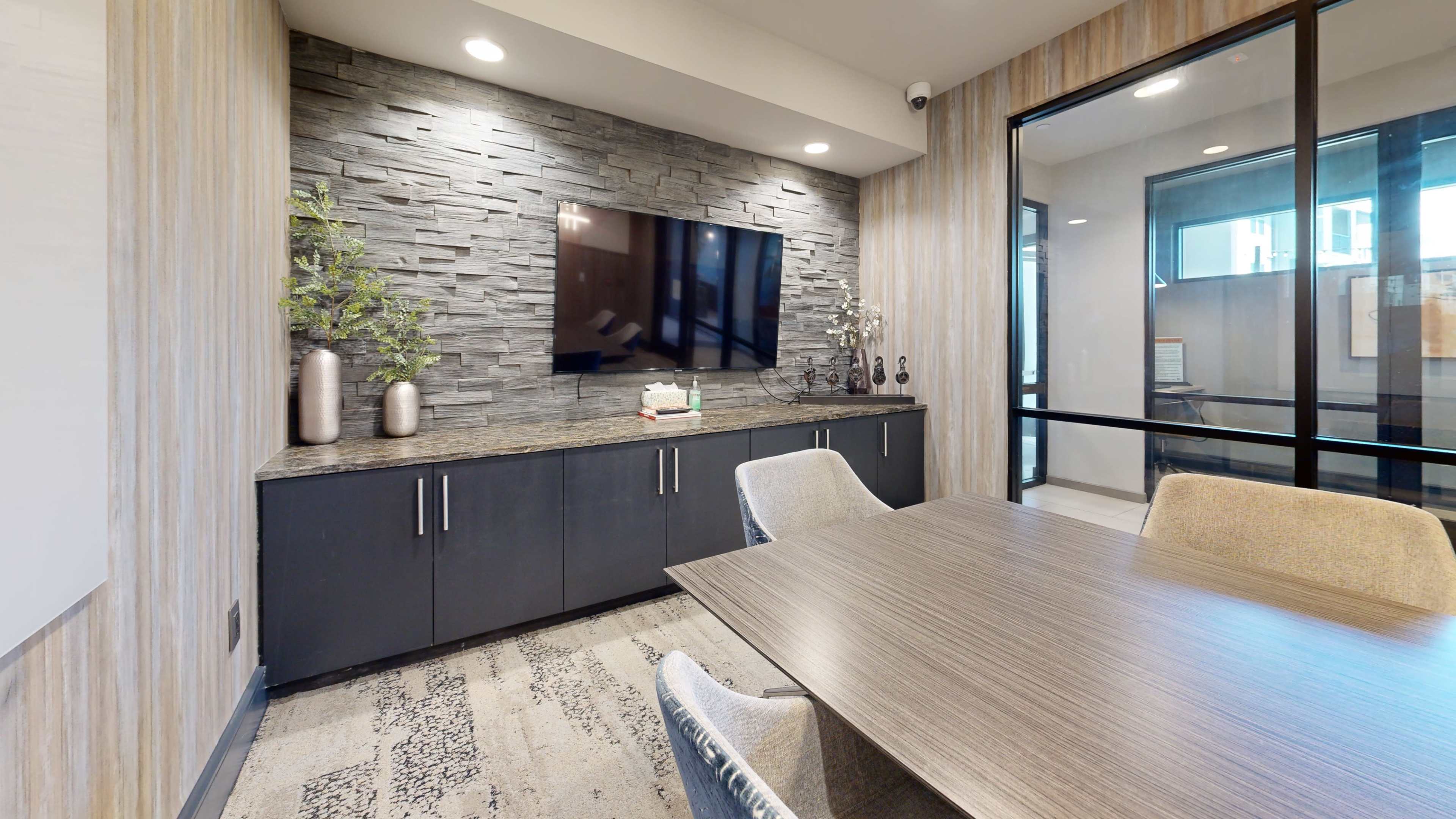 The image shows a modern conference room with a stone feature wall, a large flat-screen TV, and a wooden table surrounded by upholstered chairs.