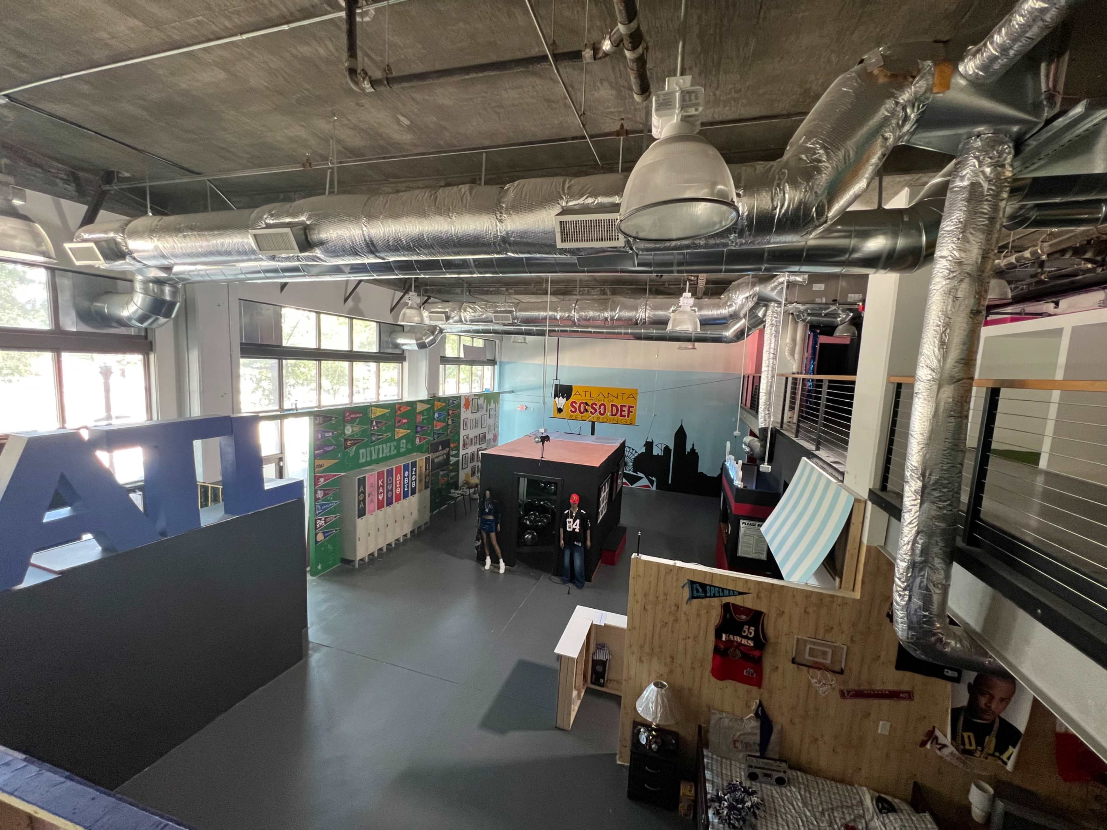 The image shows a spacious indoor area with high ceilings, featuring various colorful banners, a wooden structure, and a stage-like platform in the center.