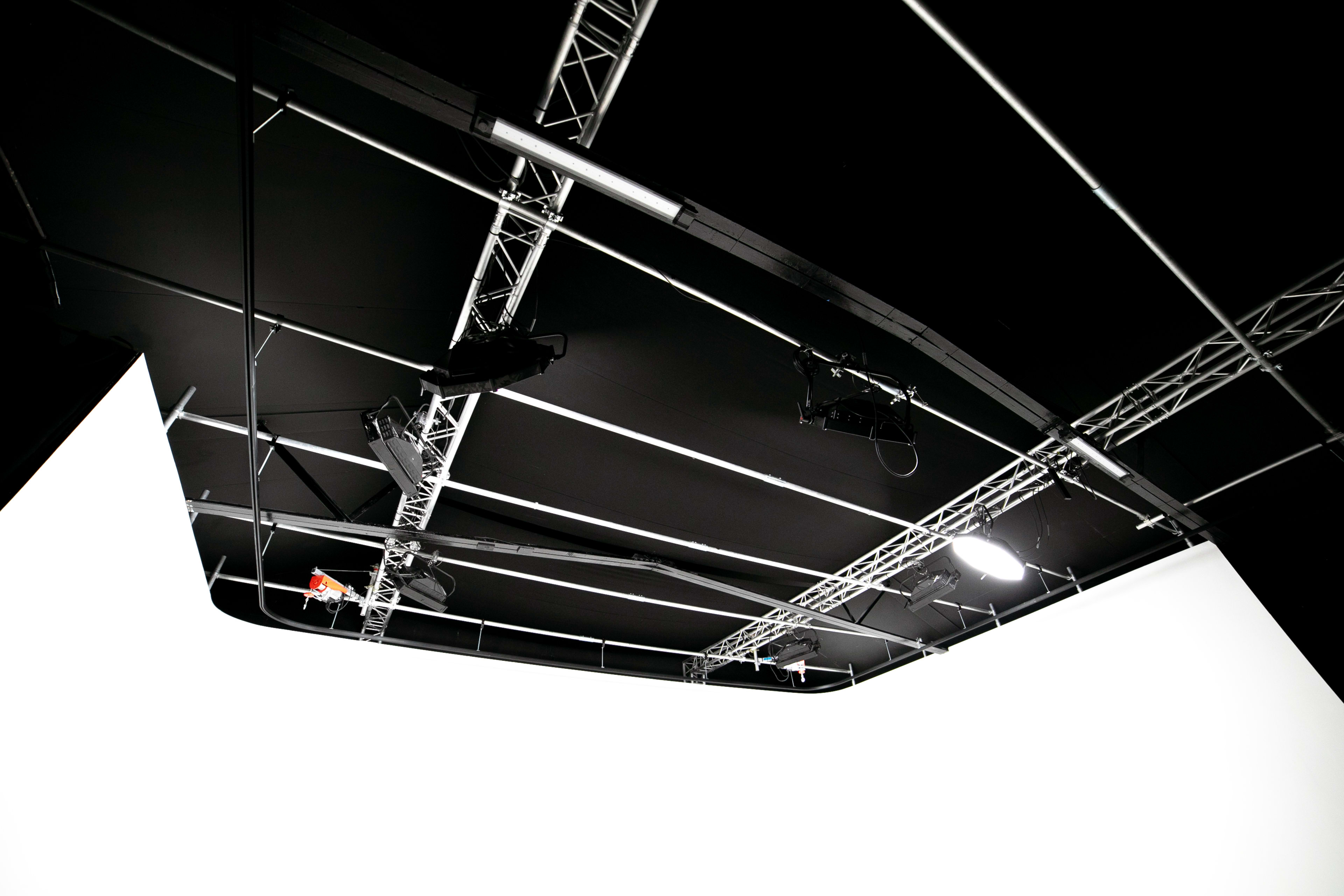 The image shows a dimly lit studio space with a white backdrop and overhead lighting equipment on a black ceiling.