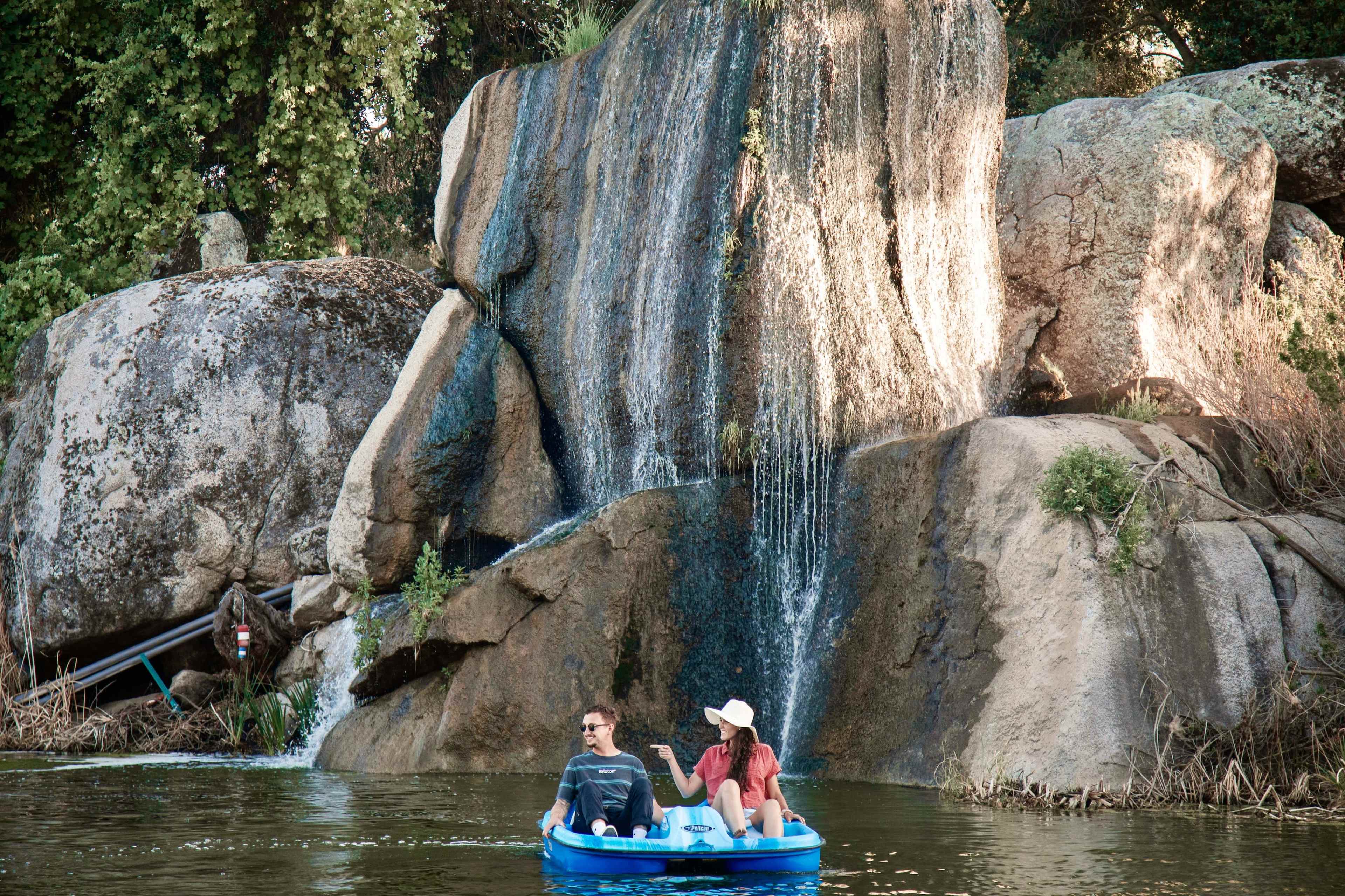 Two people are sitting in a blue inflatable boat on a body of water near a rocky cliff with a waterfall.