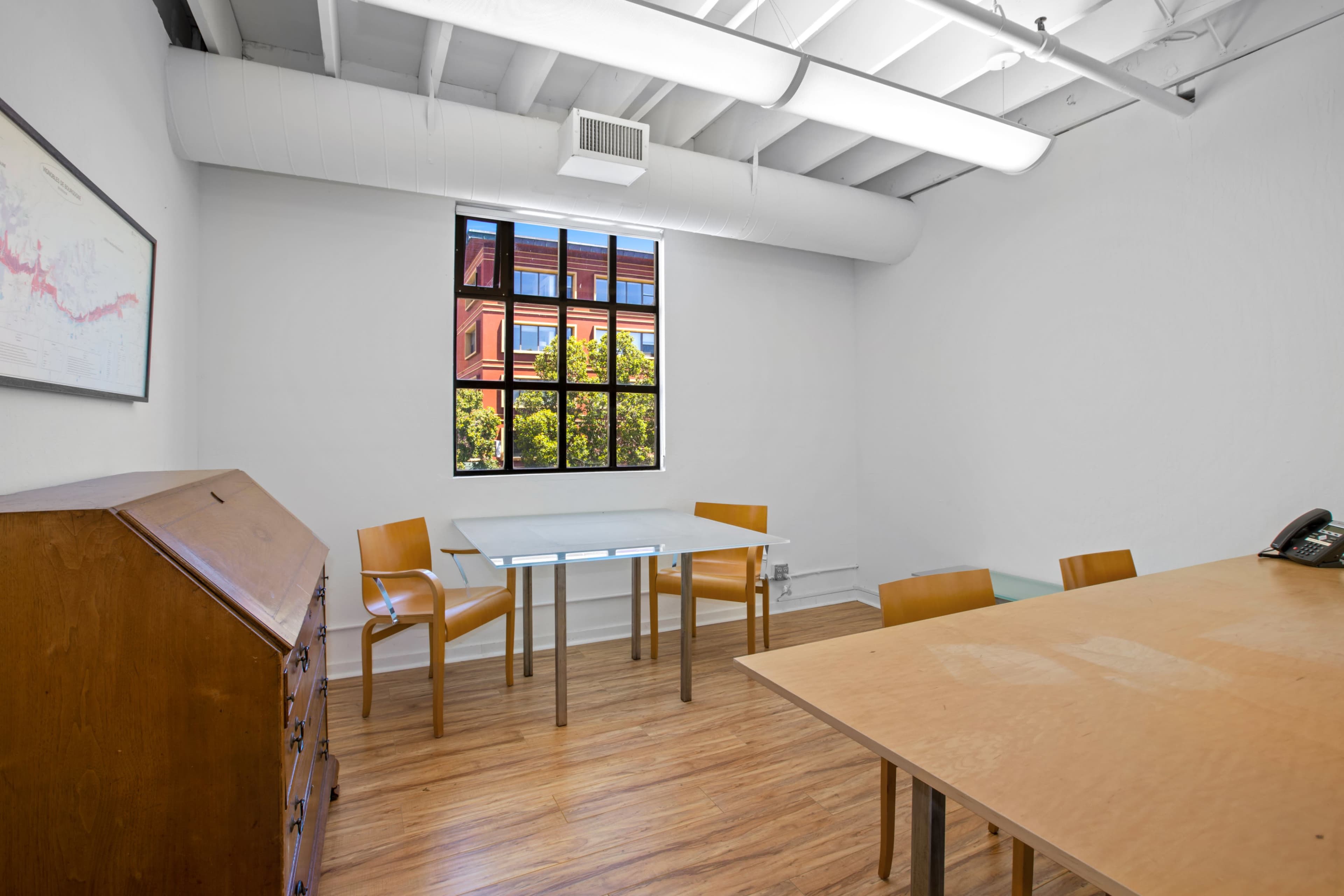 A bright, empty meeting room with a wooden table, two chairs, a telephone, and a window revealing greenery outside.