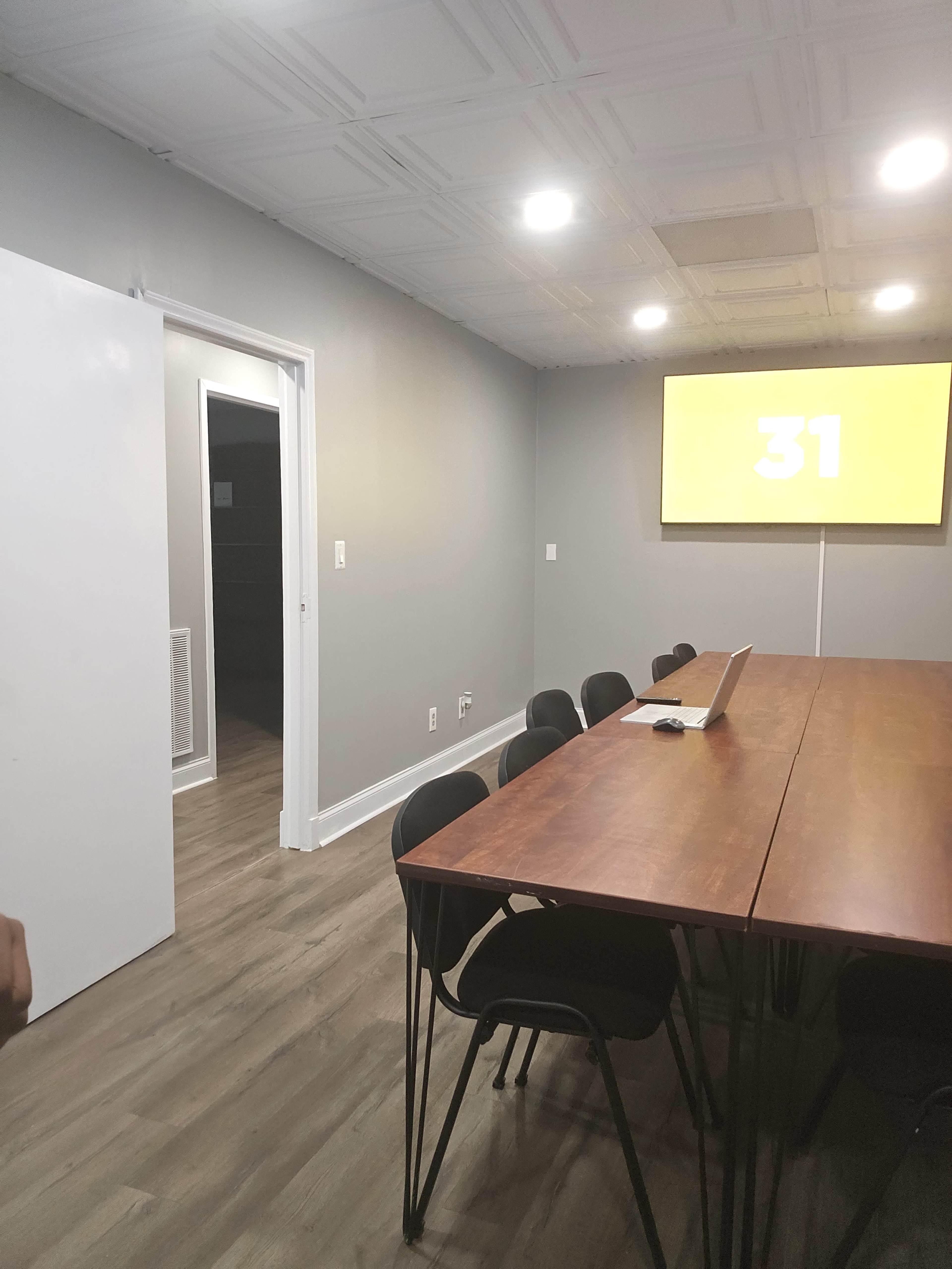 A conference room with a long wooden table, black chairs, and a large screen displaying the number 31 on a gray wall.