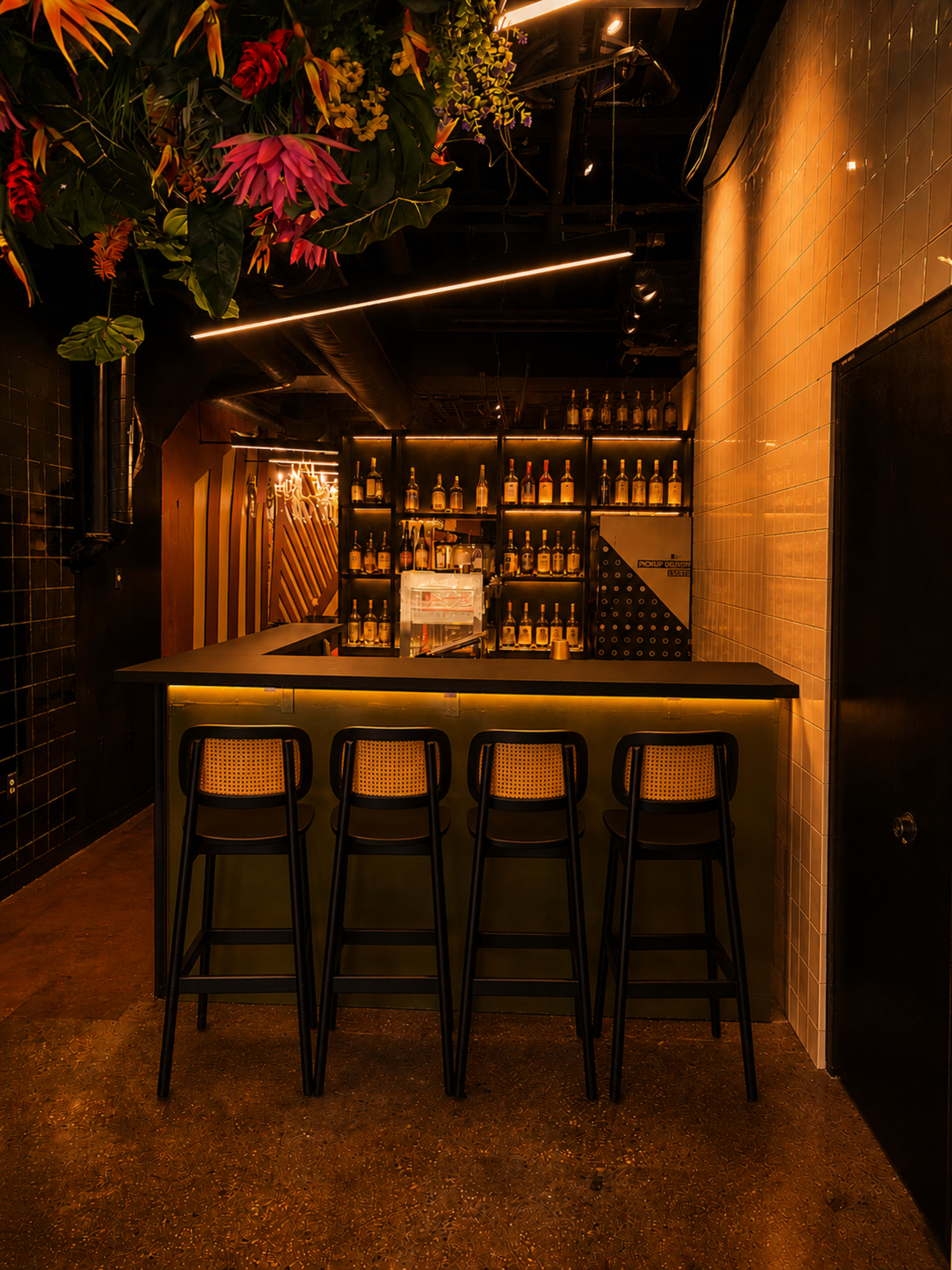 The image shows a modern bar area featuring a sleek counter with high stools, illuminated shelves displaying bottles, and decorative plants overhead.