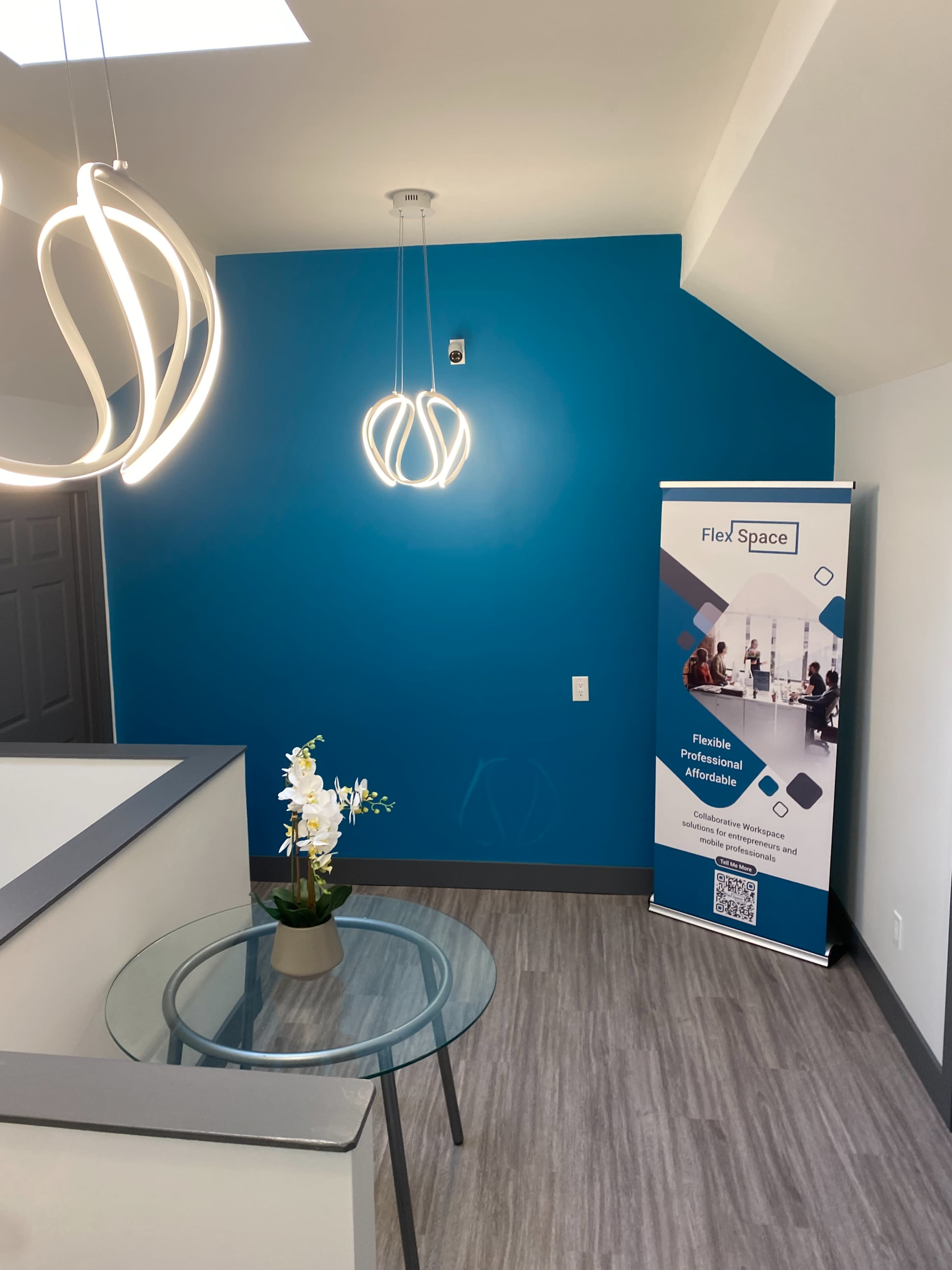 The image shows a modern office reception area with a round glass table, a potted plant, a decorative light fixture, and a banner for FlexSpace against a bright blue wall.