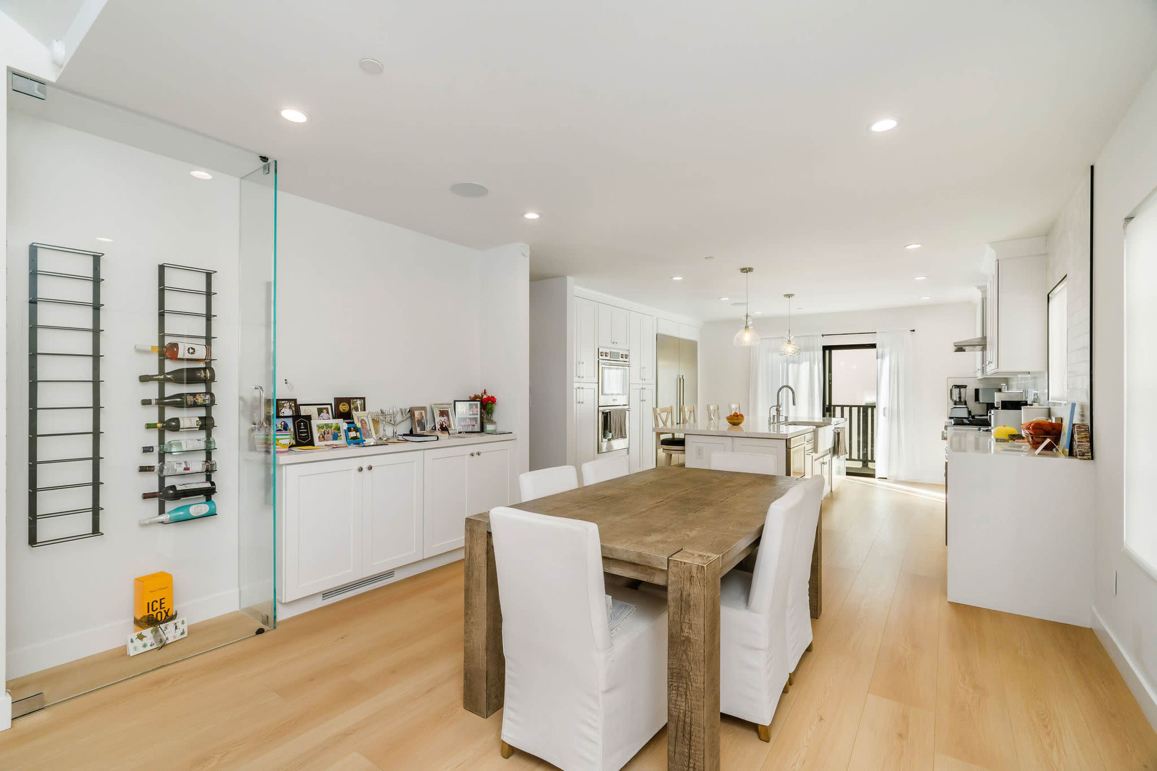 A modern kitchen and dining area features a wooden table surrounded by white chairs, with sleek cabinetry and a glass wall displaying various items.