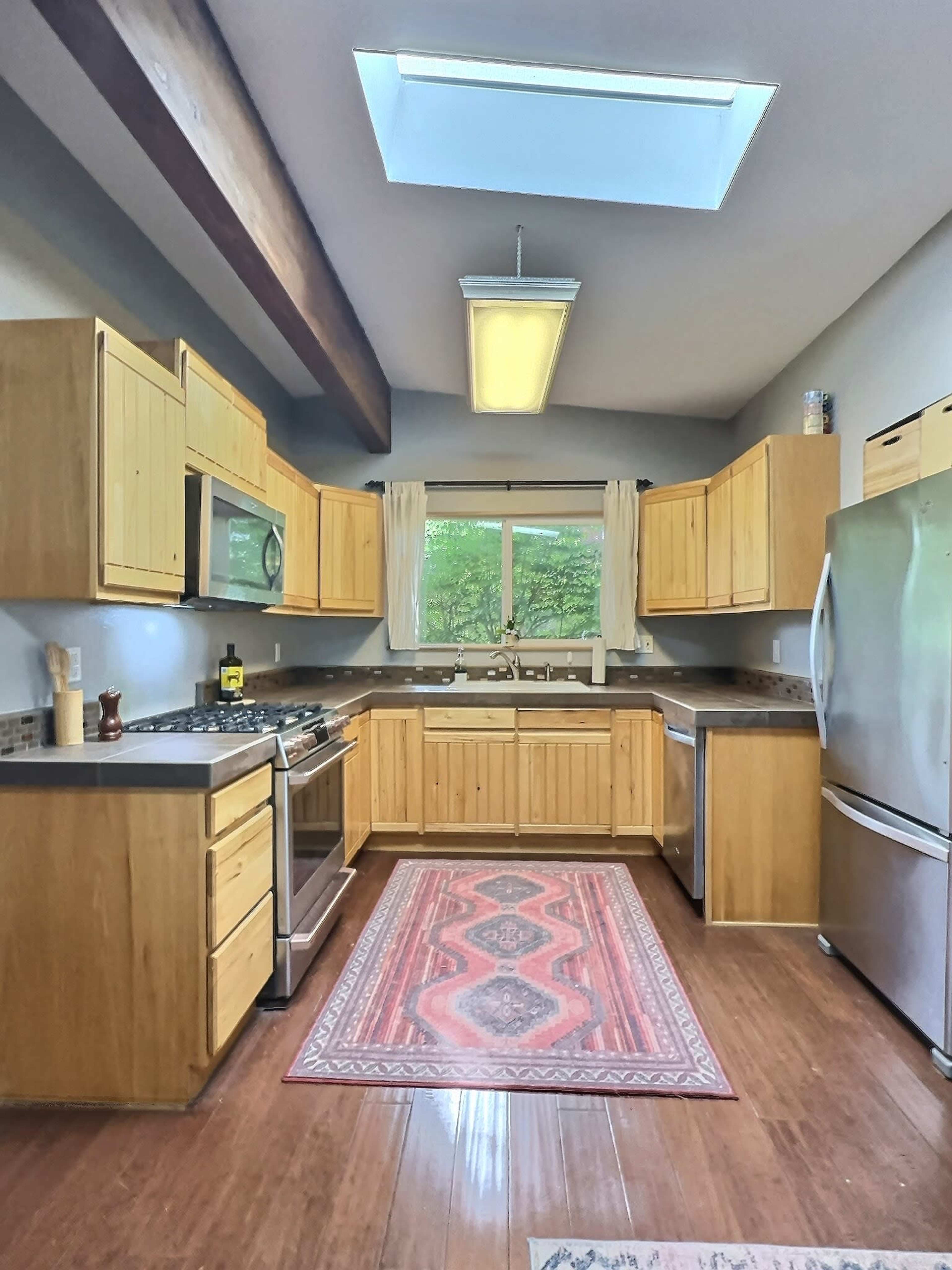 The image shows a kitchen with wooden cabinets, stainless steel appliances, a central island, and a skylight that illuminates the space.