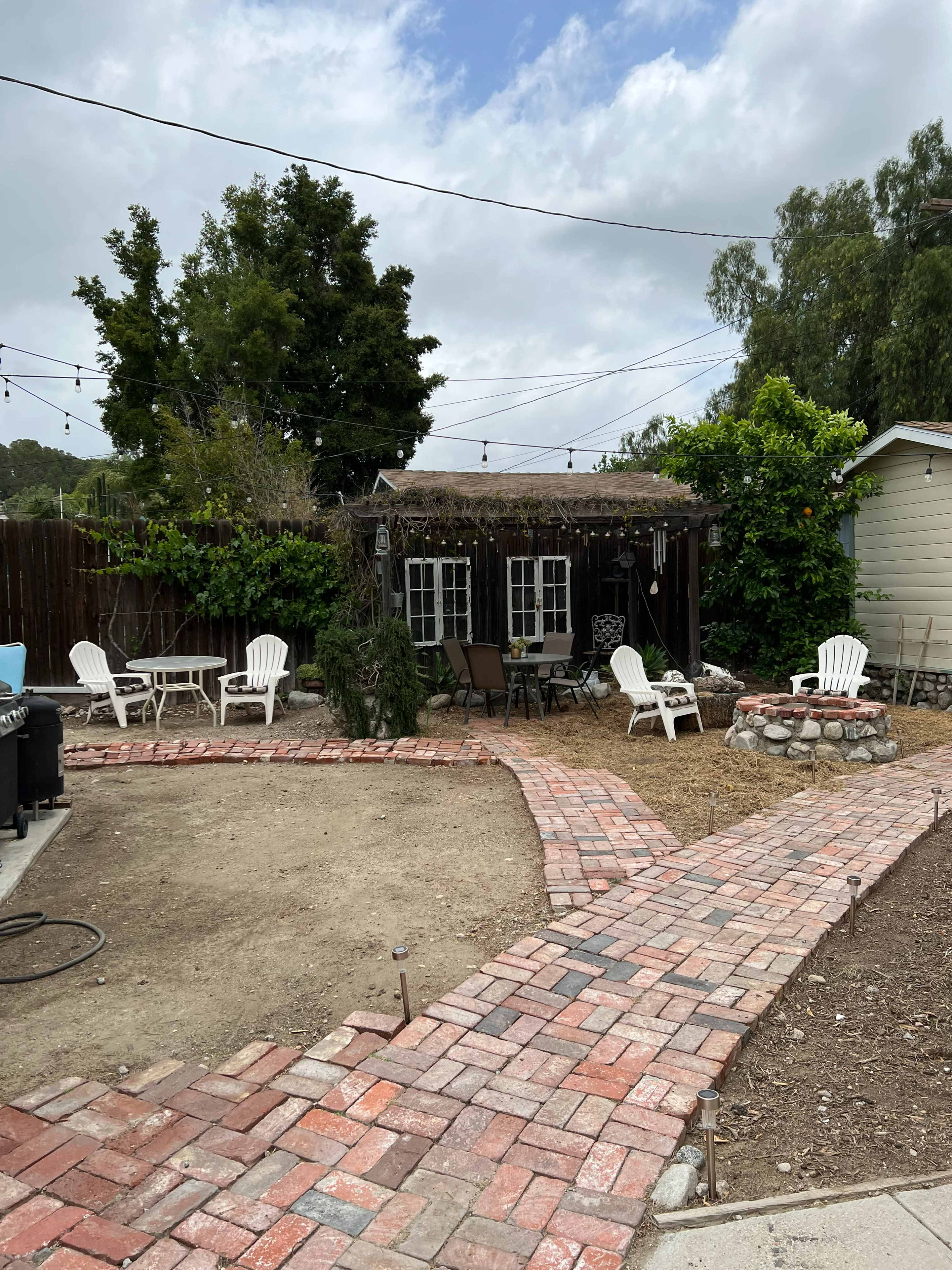The image shows a garden area with a brick pathway leading to a small outdoor seating area surrounded by trees and a shed.