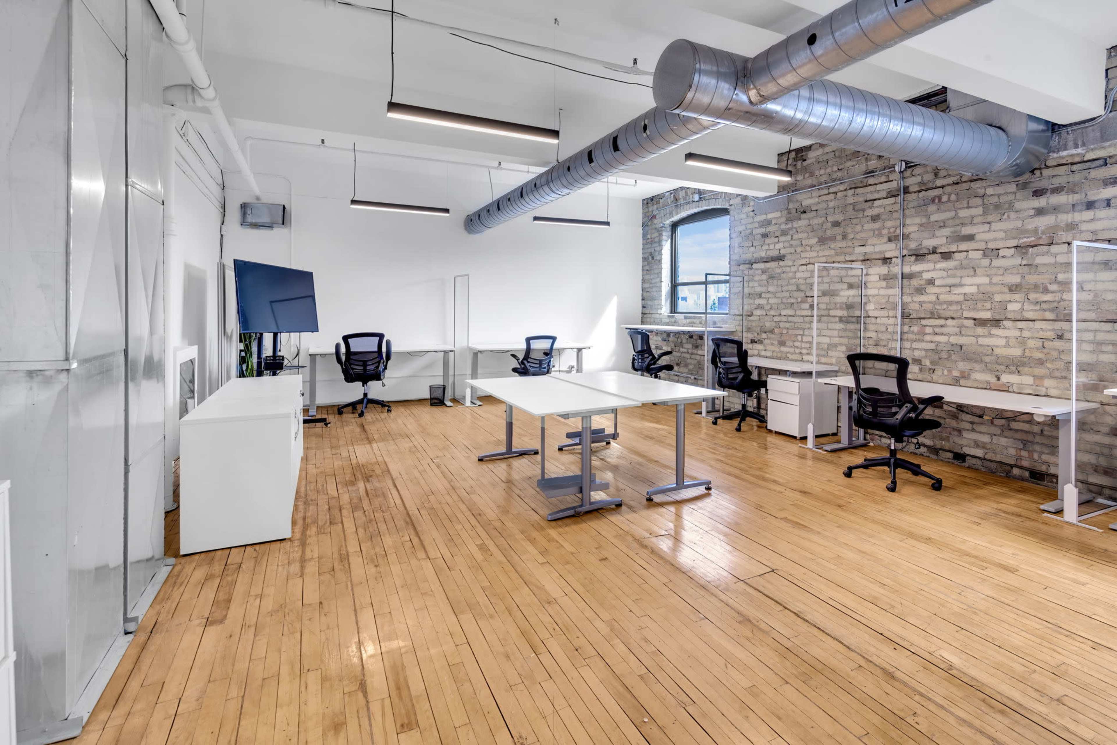 The image shows a modern office space with wooden flooring, several desks, and ergonomic chairs, featuring exposed brick walls and industrial-style lighting.