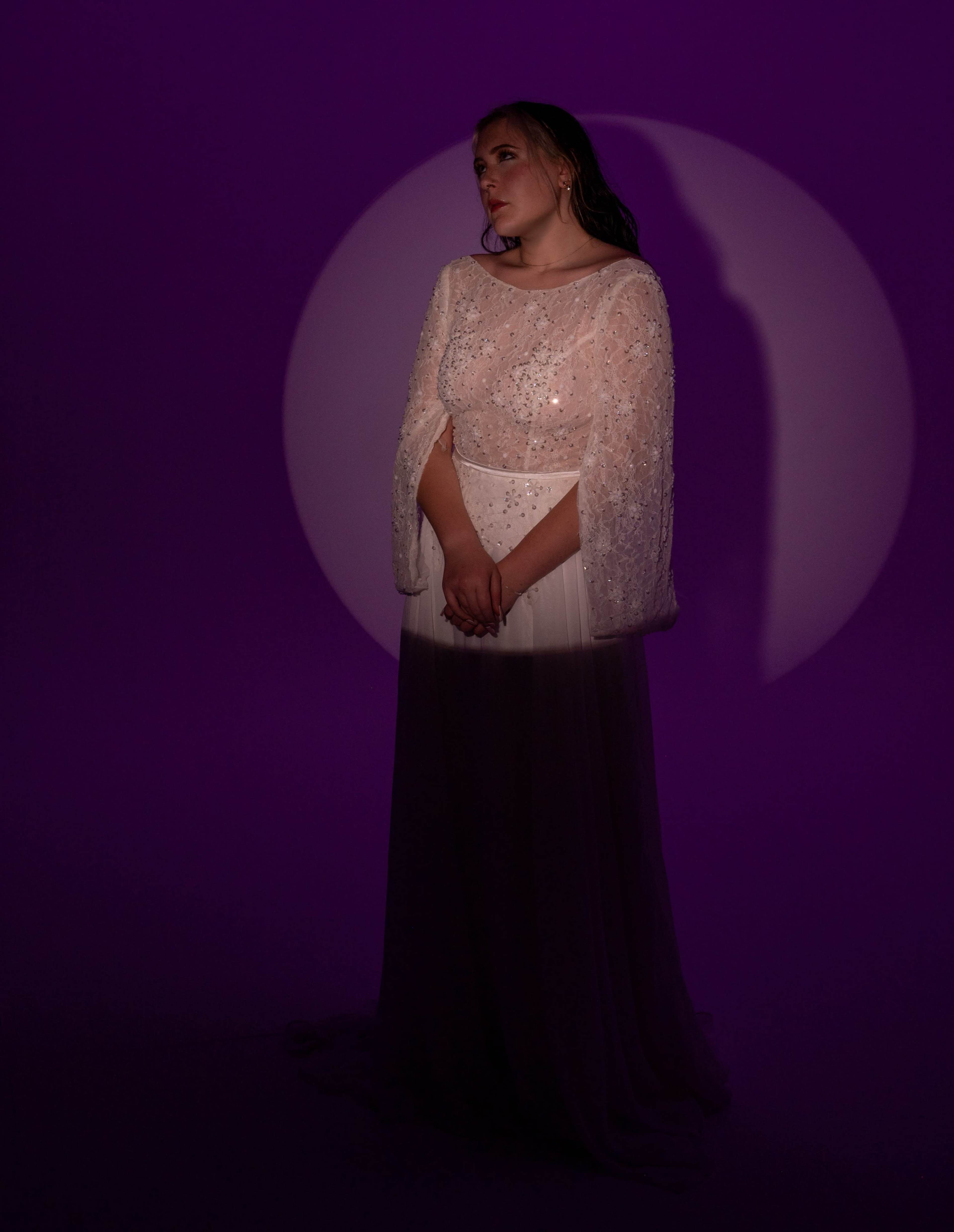 A woman in a white gown with a beaded overlay stands against a purple backdrop, illuminated by a circular light.
