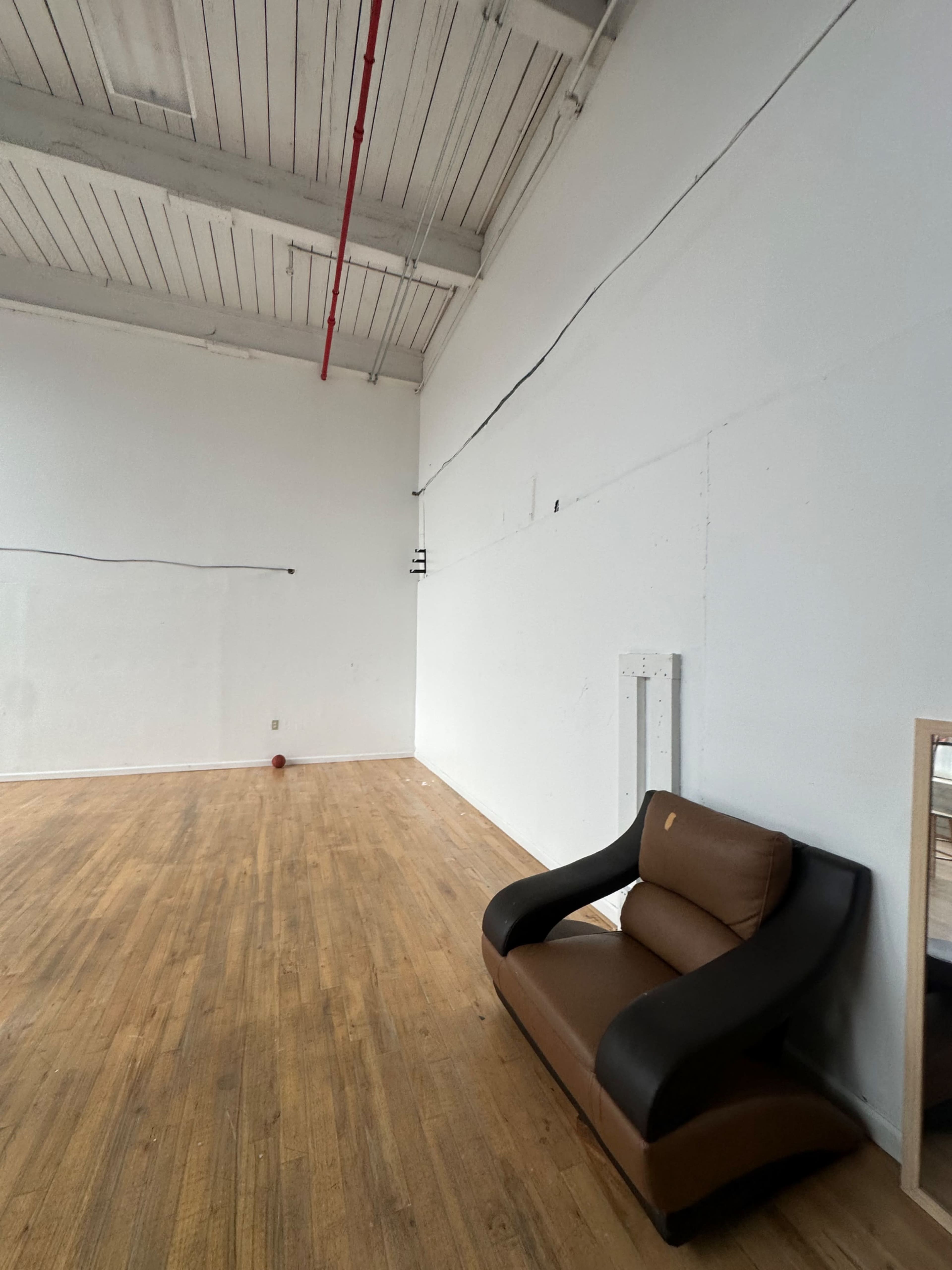 An empty room with wooden flooring features a brown and black sofa against a white wall.