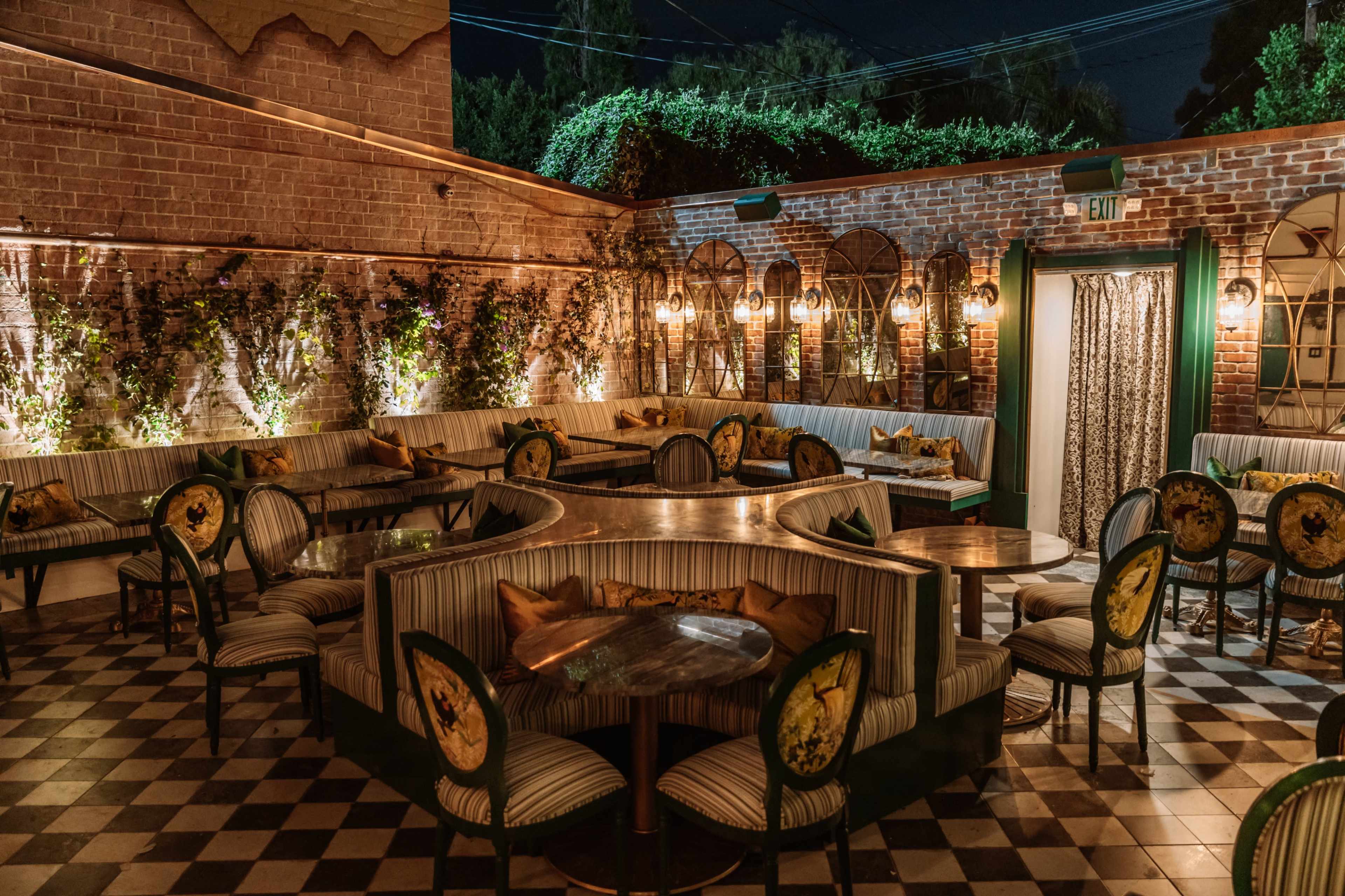 The image shows a dimly lit, elegant restaurant patio with striped seating, wooden tables, and patterned decor on the walls, surrounded by greenery.