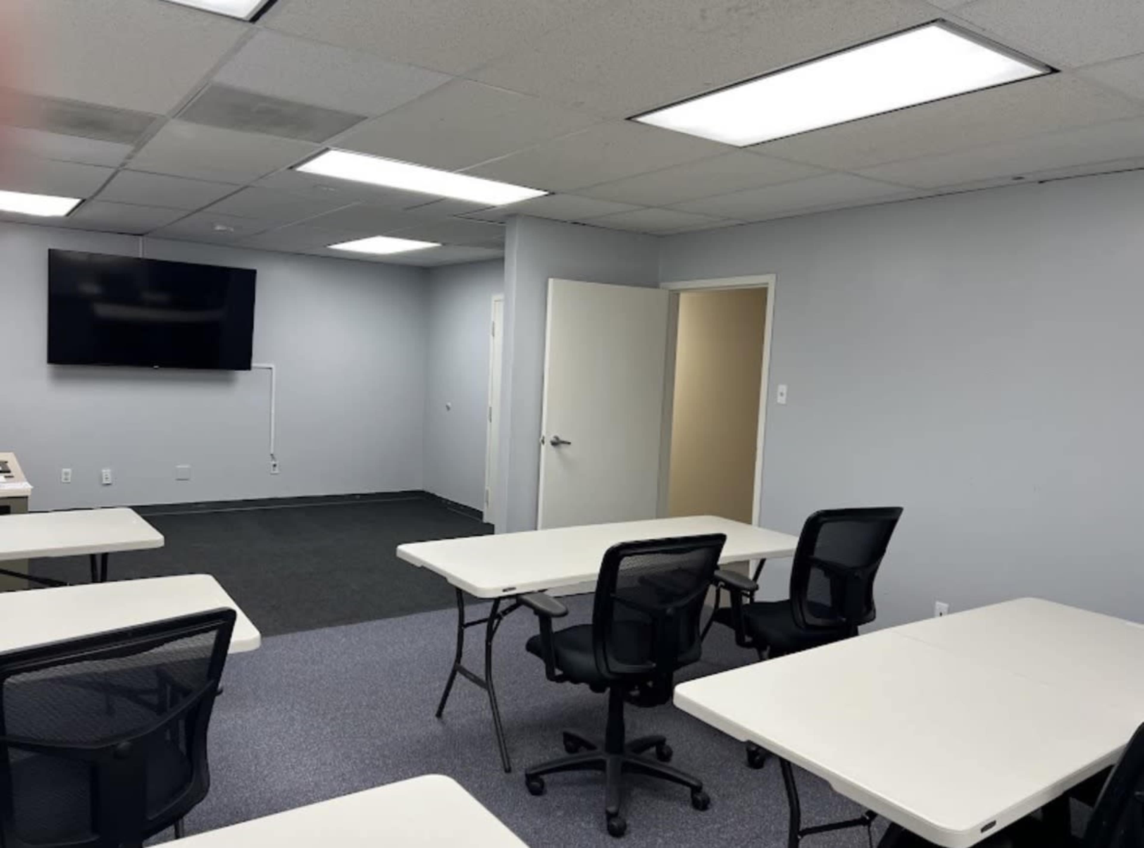 The image shows a spacious office or training room with several white tables and black chairs arranged in a semi-circle, a wall-mounted television, and a doorway leading to another room.