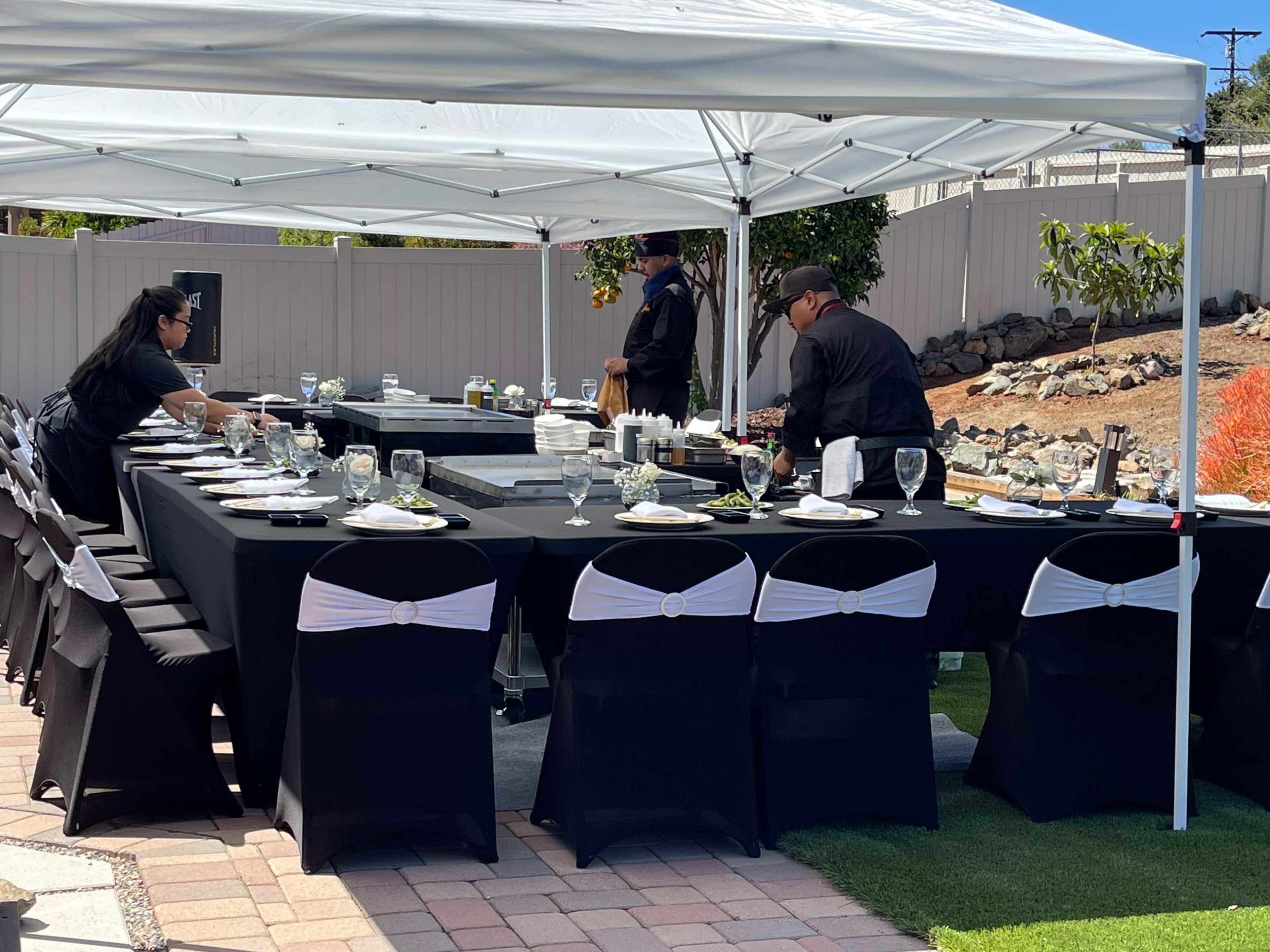 A dining setup is arranged under a tent with black tablecloths, white accents on the chairs, and staff preparing the table.